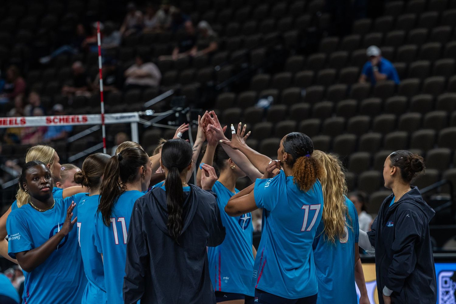 Women's volleyball team in teal uniforms huddle together raising hands in a team cheer before a match.