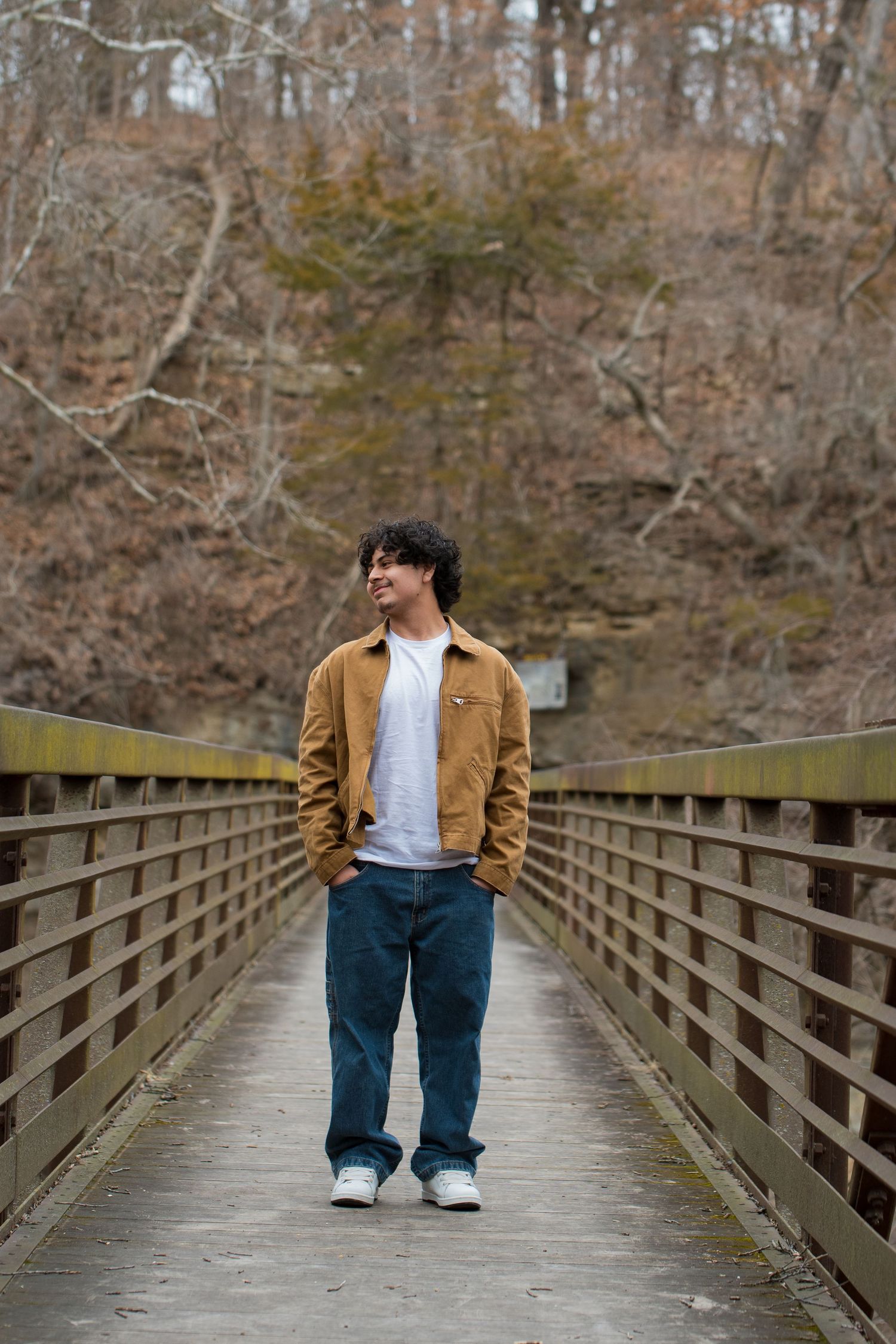 Young man in brown jacket and jeans stands on wooden bridge surrounded by autumn trees.