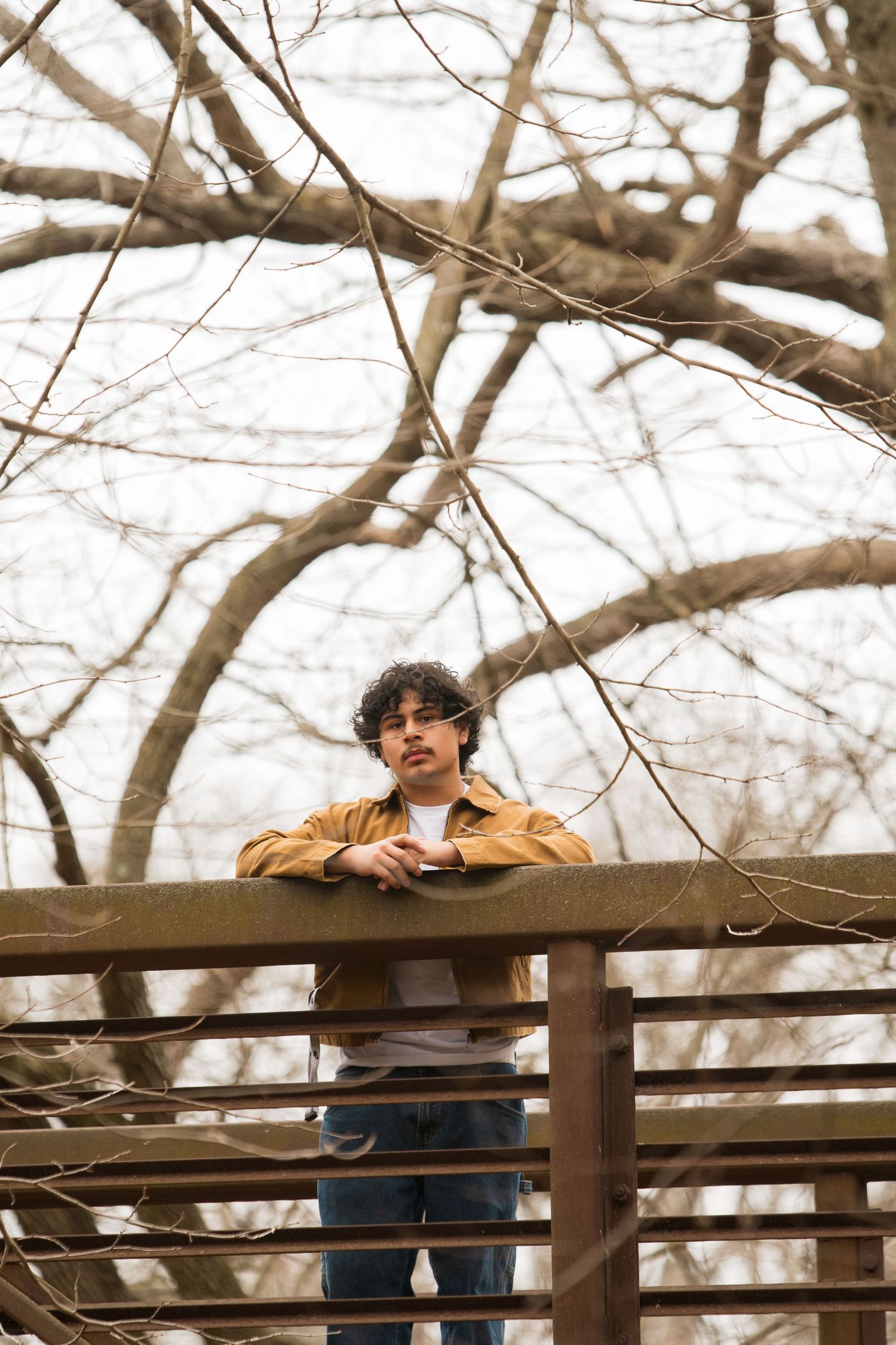 Young man in yellow jacket leaning on wooden railing surrounded by bare tree branches on overcast day.