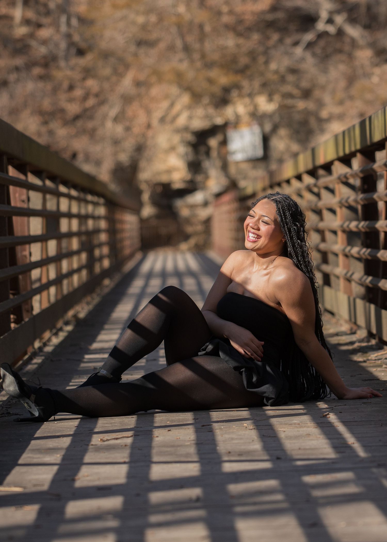 A smiling woman in black outfit and patterned tights sits on a wooden bridge in a sunny outdoor setting.