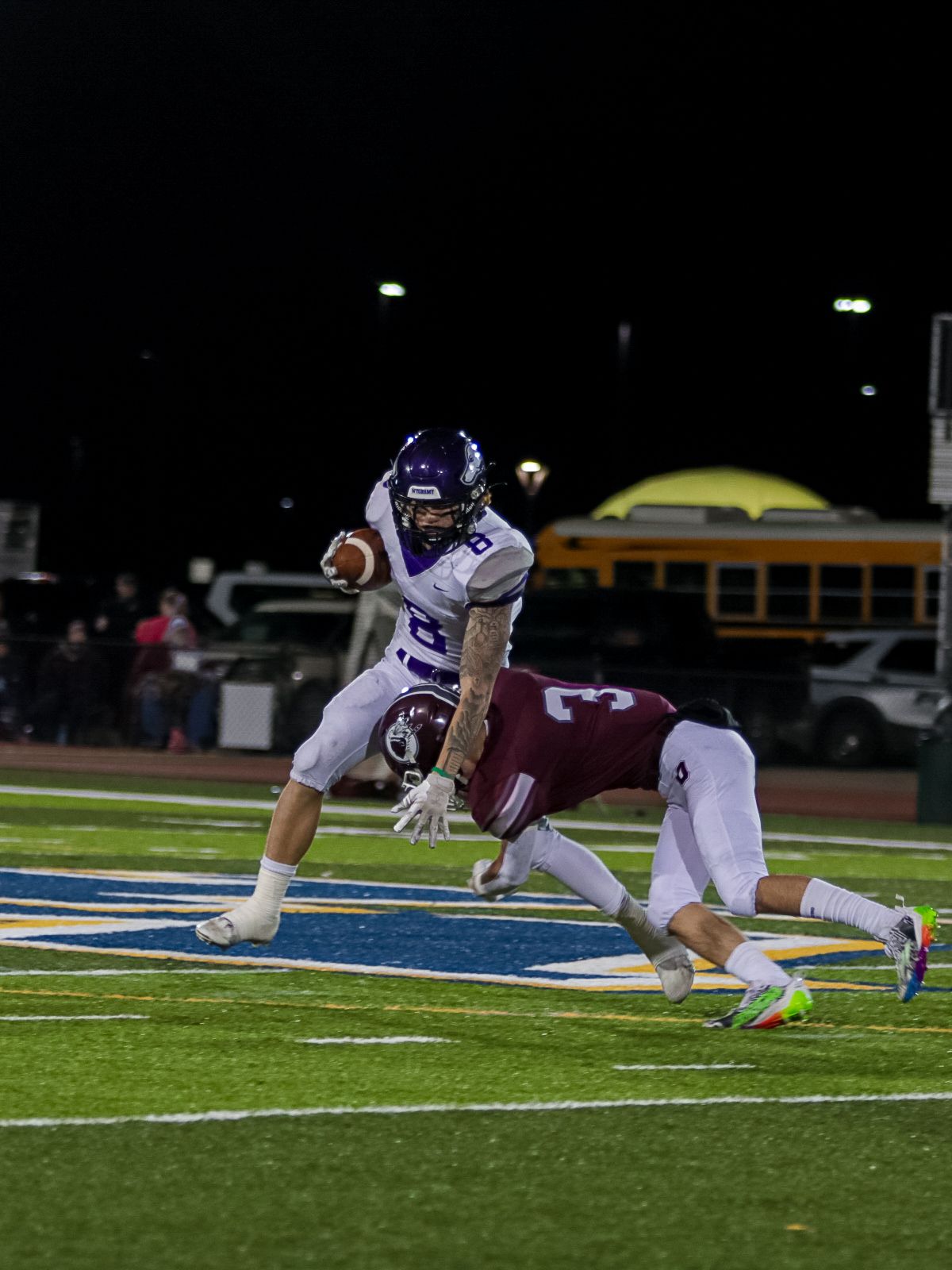 Two football players collide during a nighttime game as one attempts to tackle the other on a lit field.