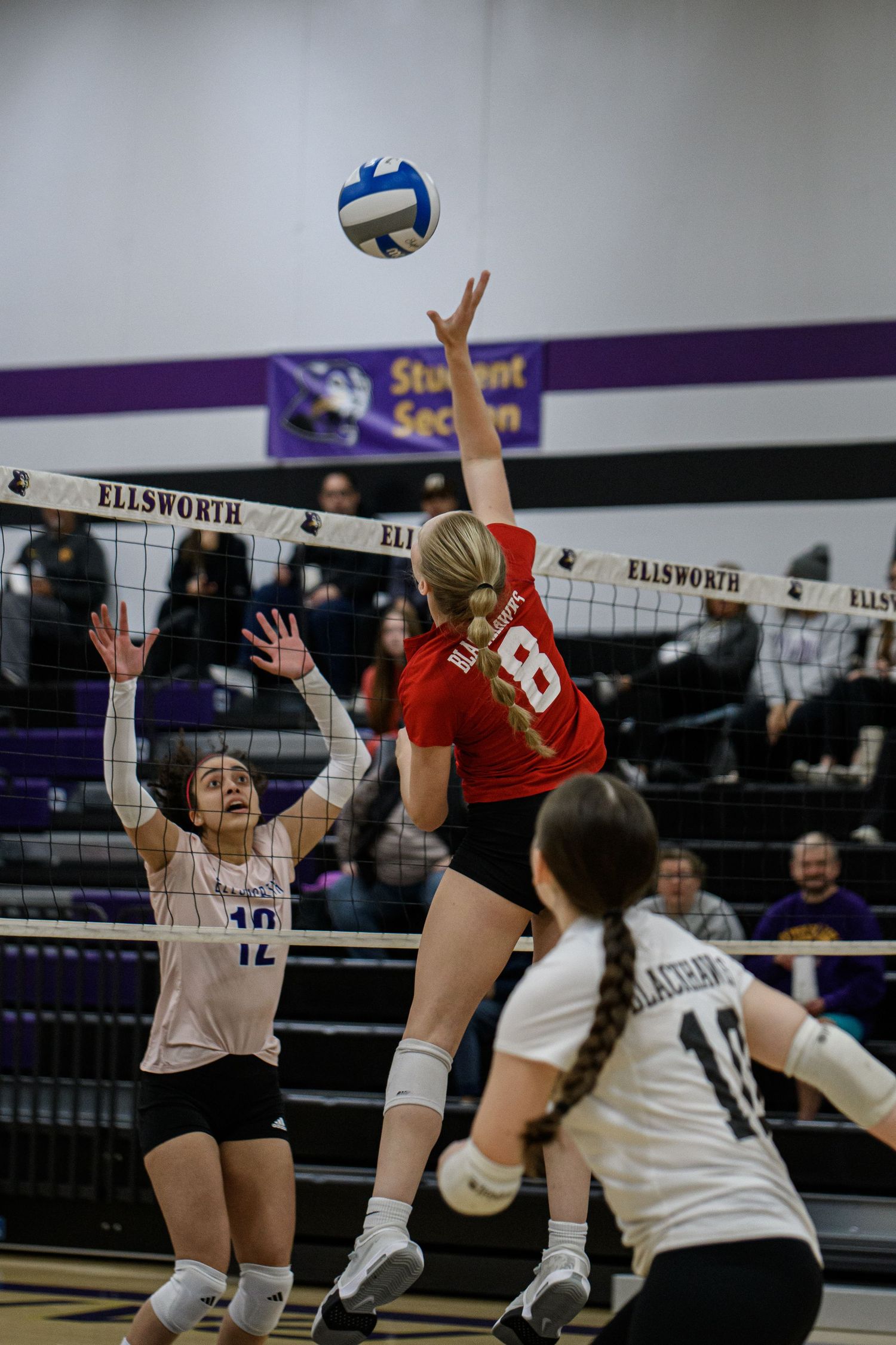 High school volleyball player in red jersey #8 spikes ball over net at Ellsworth gym during competitive match.