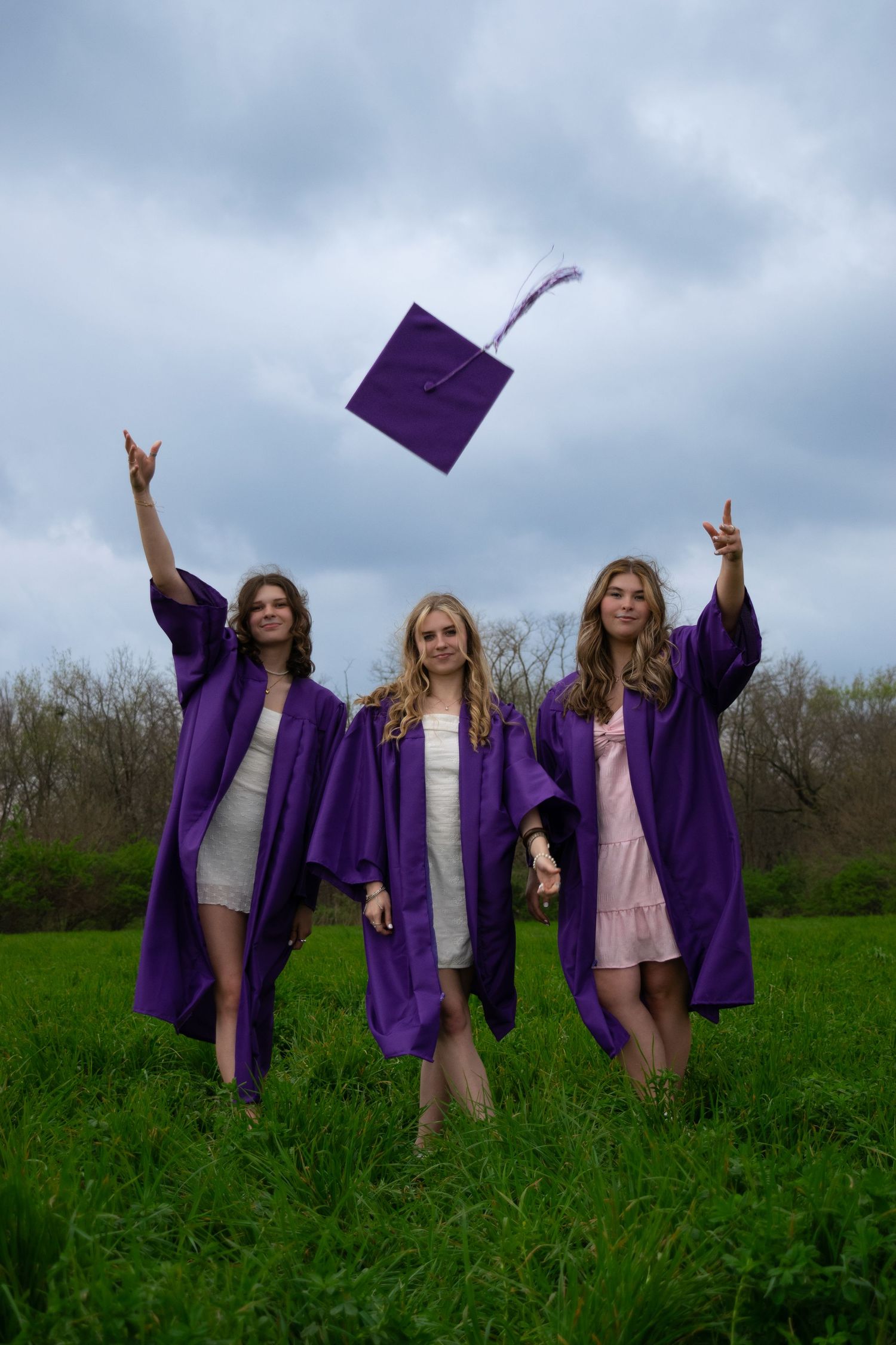 Graduates in purple caps and gowns celebrate together in a green field under cloudy skies.