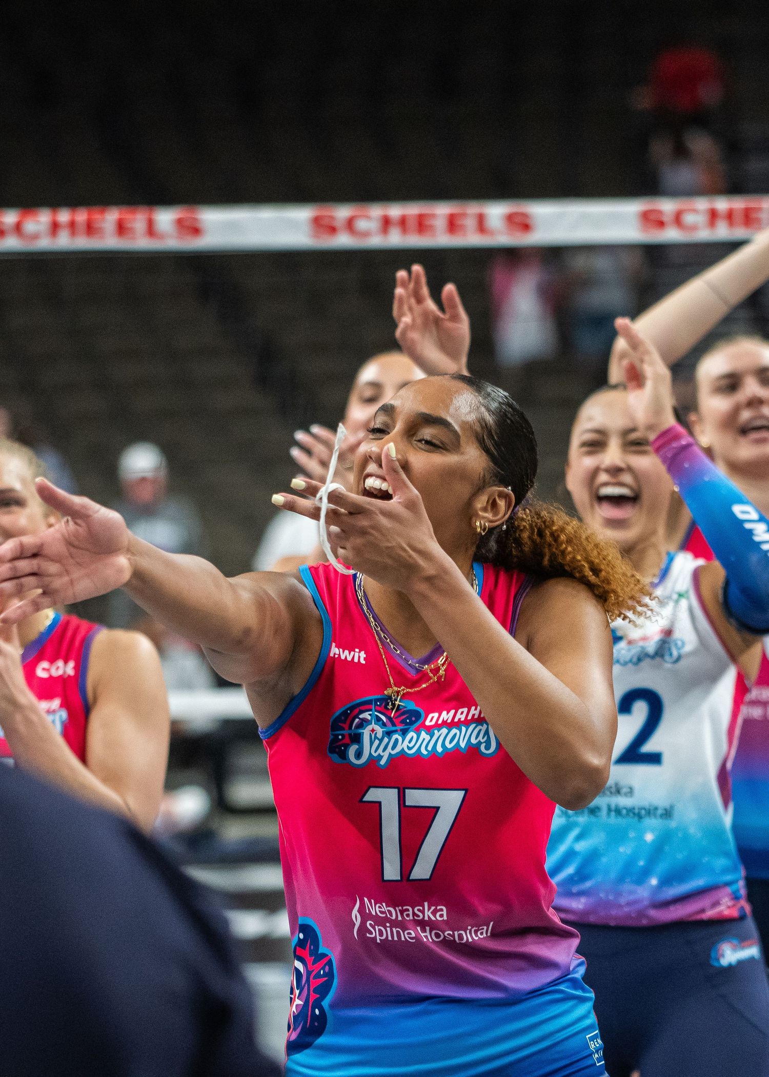 Omaha Supernovas player #17 celebrates with teammates in pink and blue uniforms at a SCHEELS volleyball event.