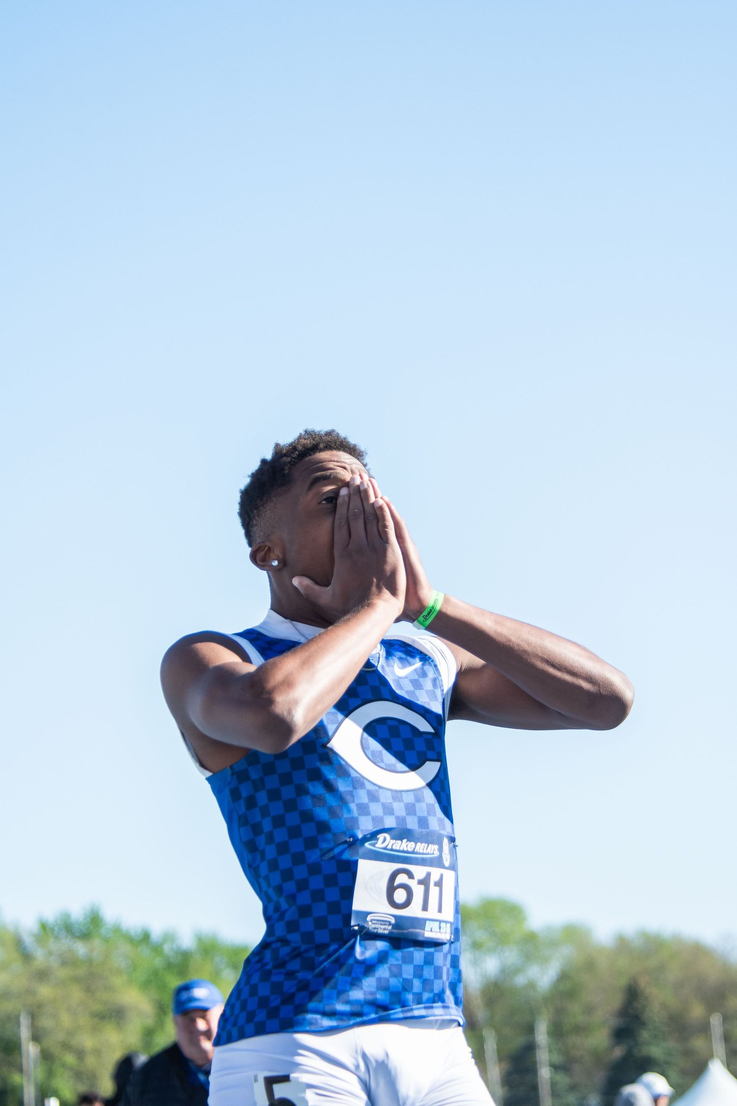 Track athlete #611 in blue checkered jersey covers face with hands at outdoor competition on sunny day.