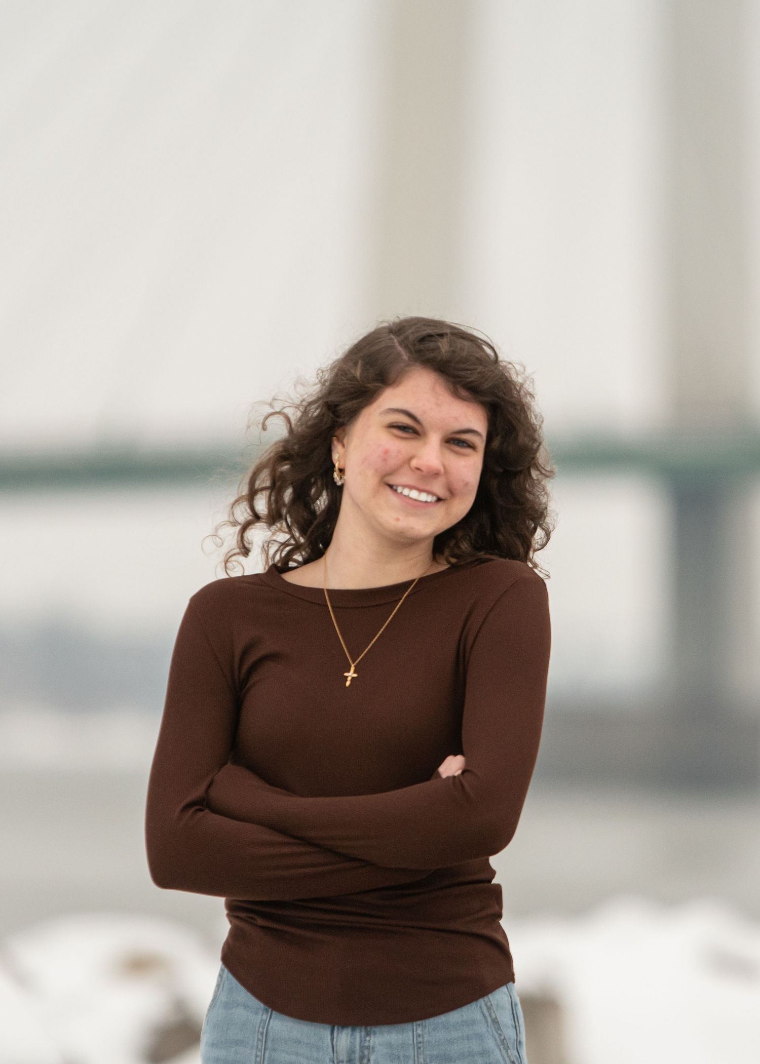 Young woman with curly hair smiling, wearing brown top and gold cross necklace, arms crossed outdoors.