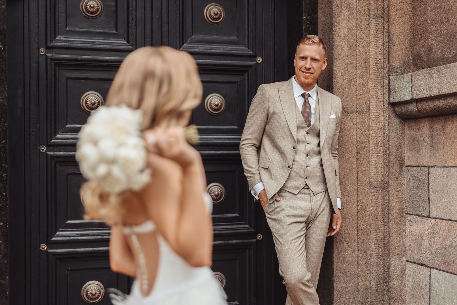 Vestuvių fotografija, kurioje nuotaka iš tolo žvelgia į savo vyrą, kuris stovi atsirėmęs į Lietuvos Banko pagrindines duris