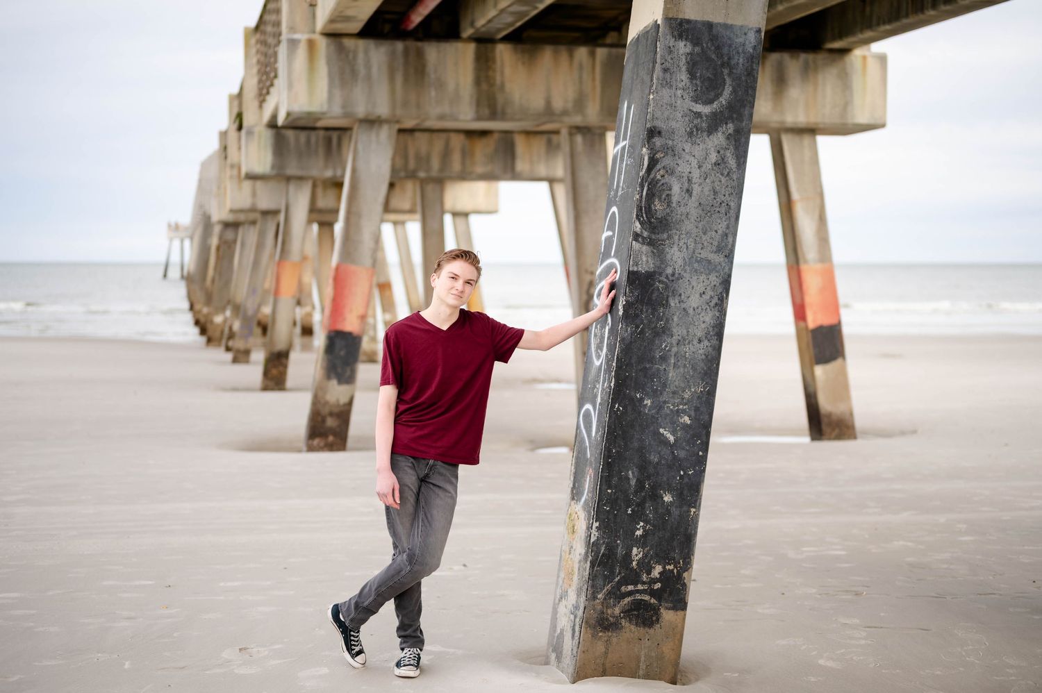 Person in red sweater walking by weathered concrete pillars below a pier at sunset.
