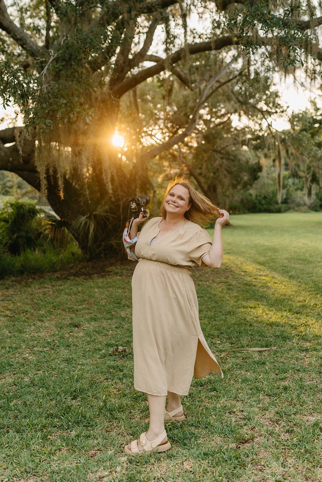 Jessica Life Photography owner in flowy beige dress twirls in grassy field during golden hour with trees in background.