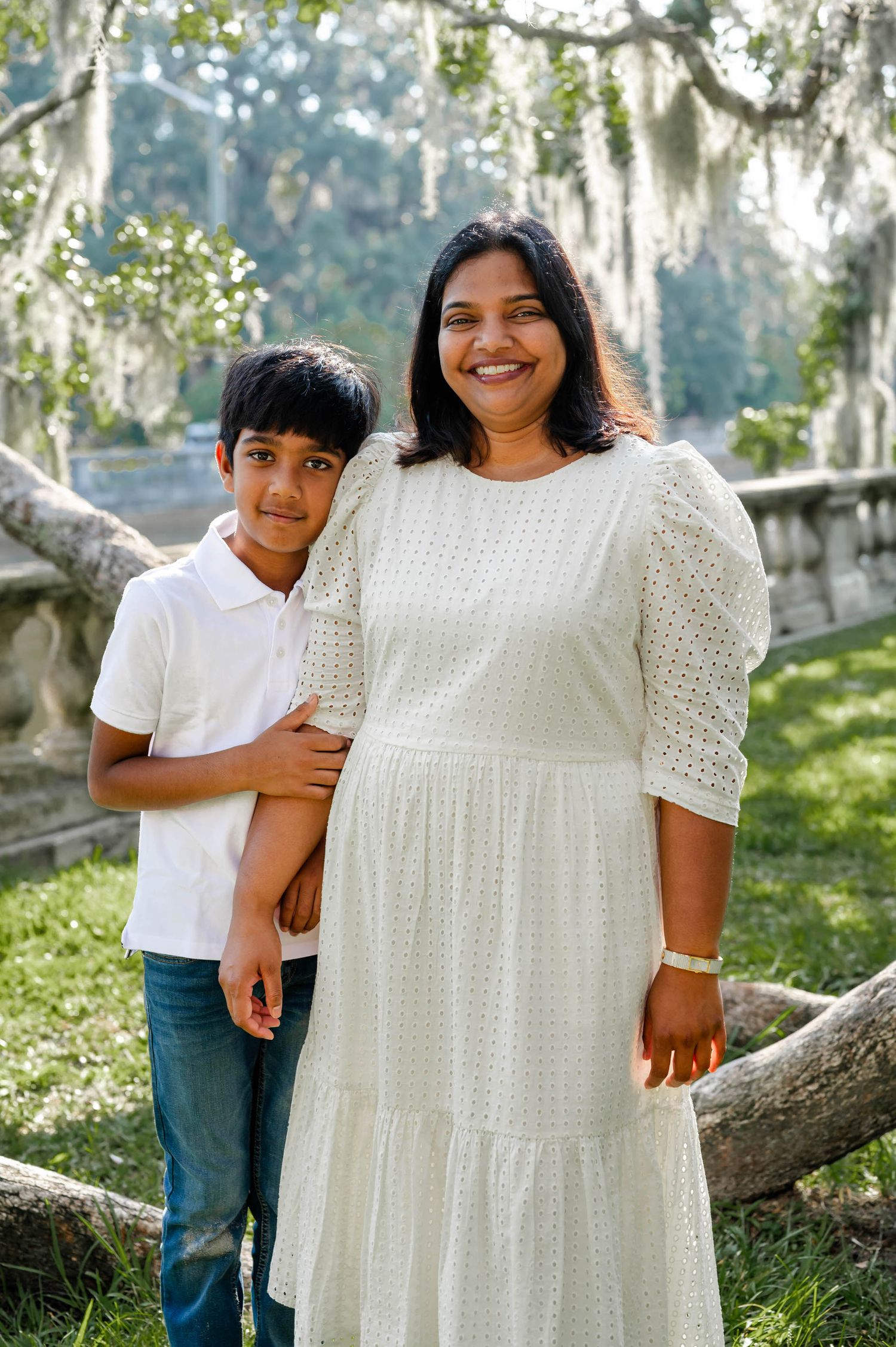 A pair in white outfits pose together outdoors with Spanish moss and trees in the background.