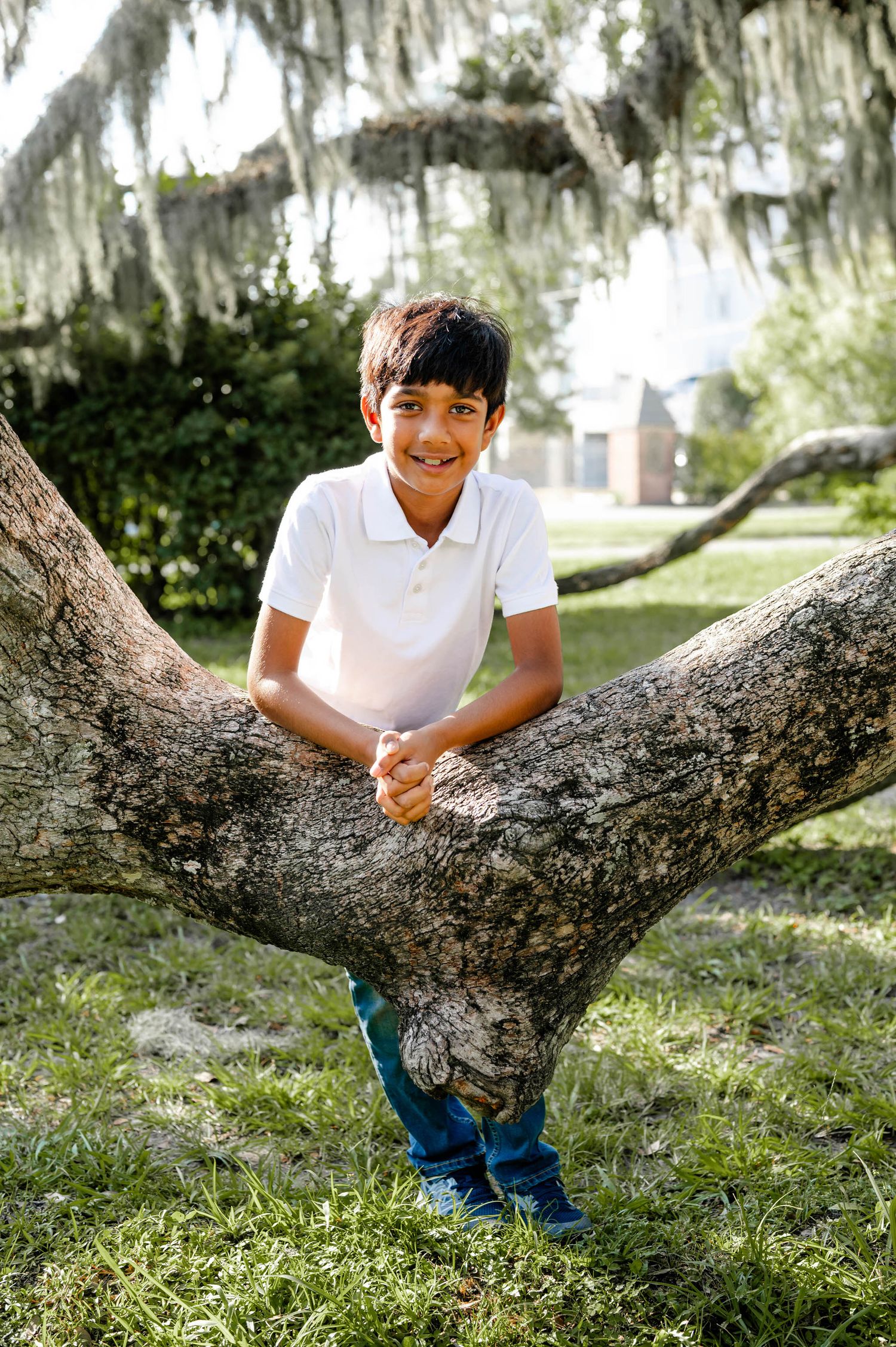 Person in white polo shirt and blue pants sits playfully on a large tree branch under Spanish moss.