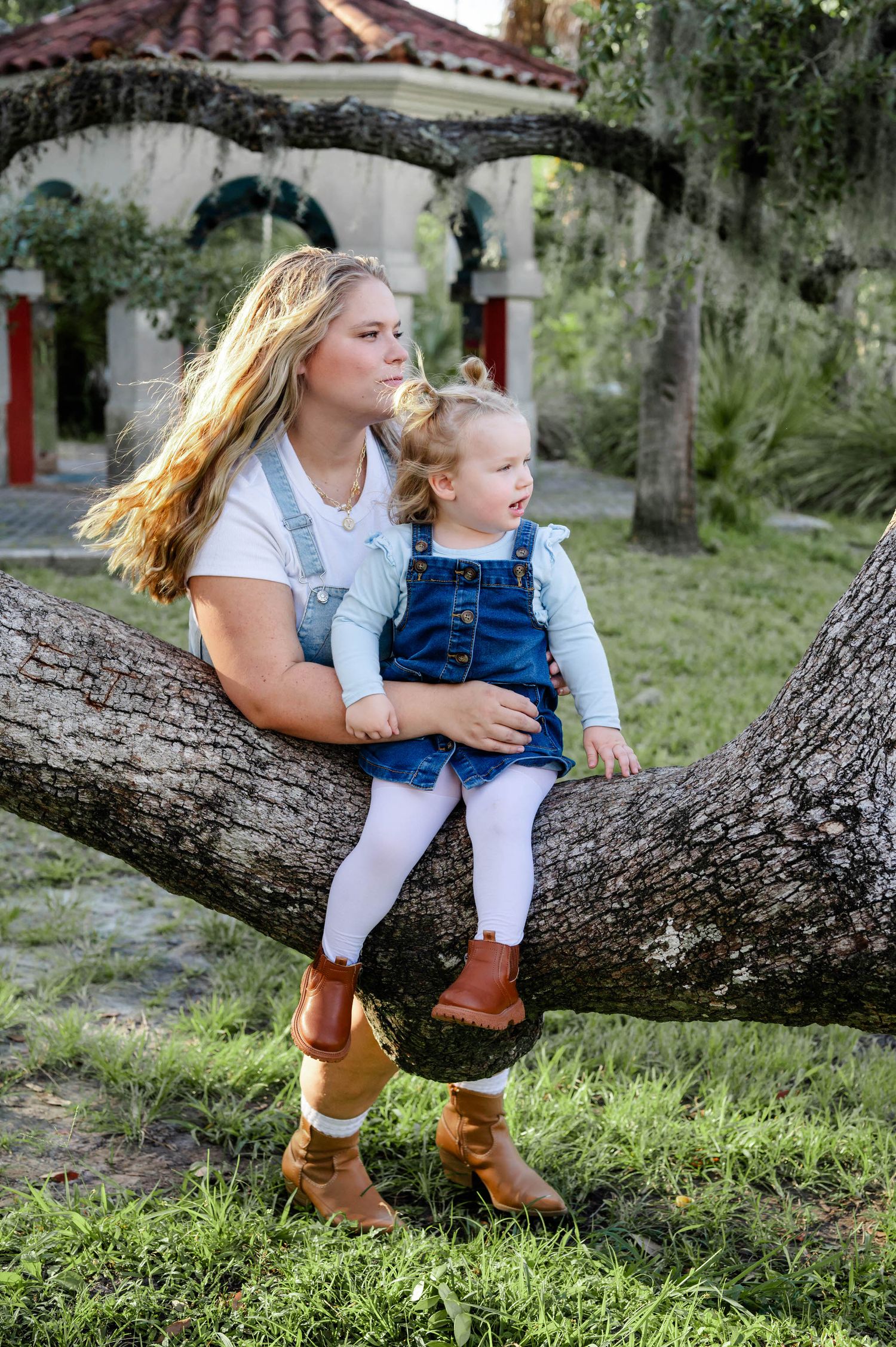 A parent and child sit together on a large tree branch, wearing casual clothing and boots in an outdoor garden setting.