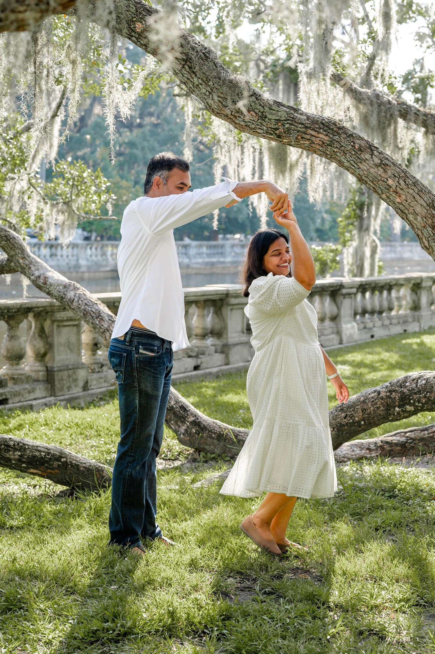 A couple dances playfully together in a sunlit garden with trees and stone balustrades in the background.