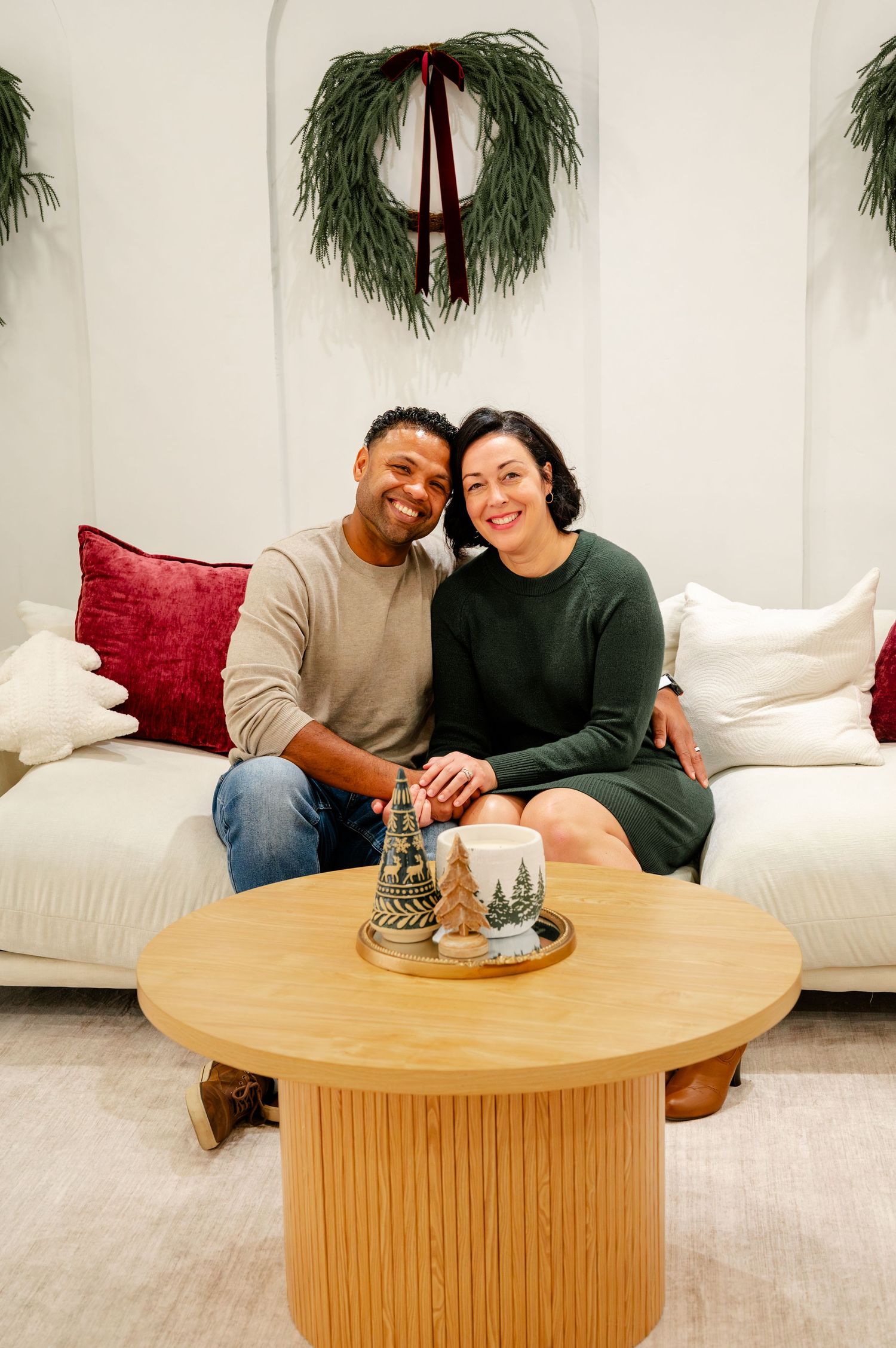 A couple sits together on a white couch with holiday decor and a round wooden coffee table in their cozy living room.