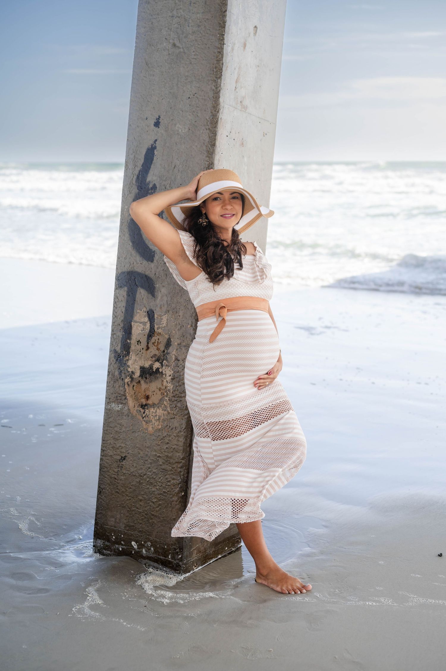Maternity photoshoot at beach with woman in white striped dress posing against concrete pillar.