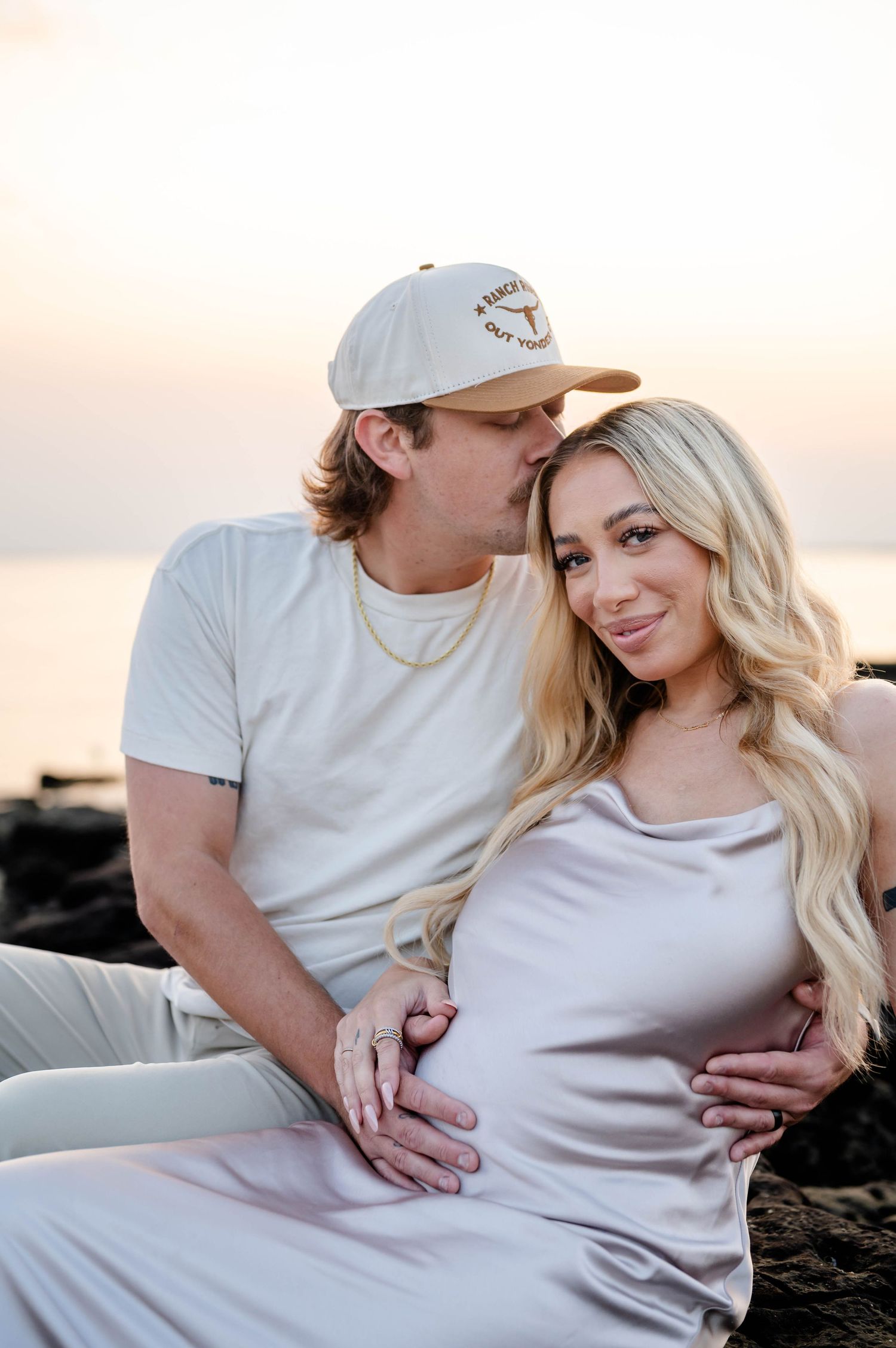 A tender maternity beach photo session at sunset with couple in white and beige clothing.