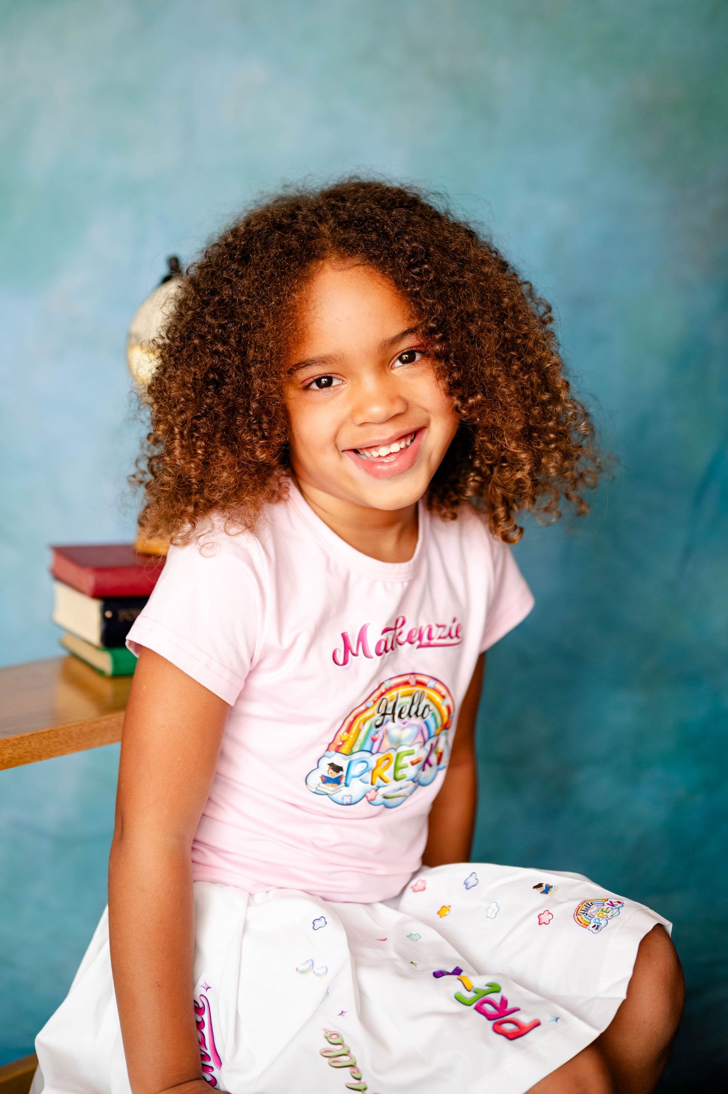 Girl in pink shirt smiling and sitting against turquoise backdrop by a Jacksonville preschool photographer.