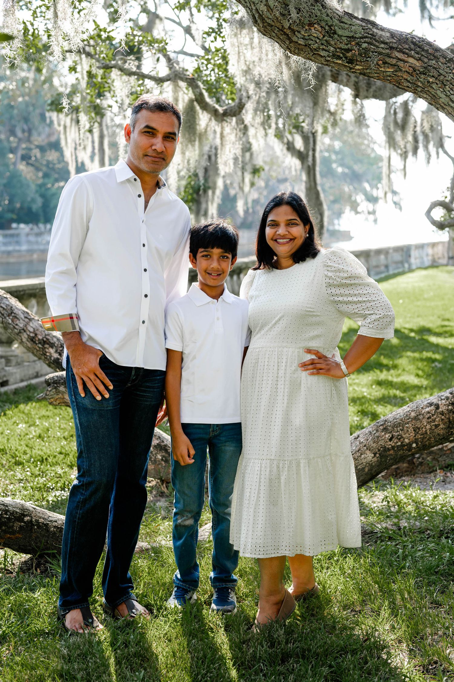 A family of three poses together under a Spanish moss-draped tree on a sunny day wearing coordinated white outfits.