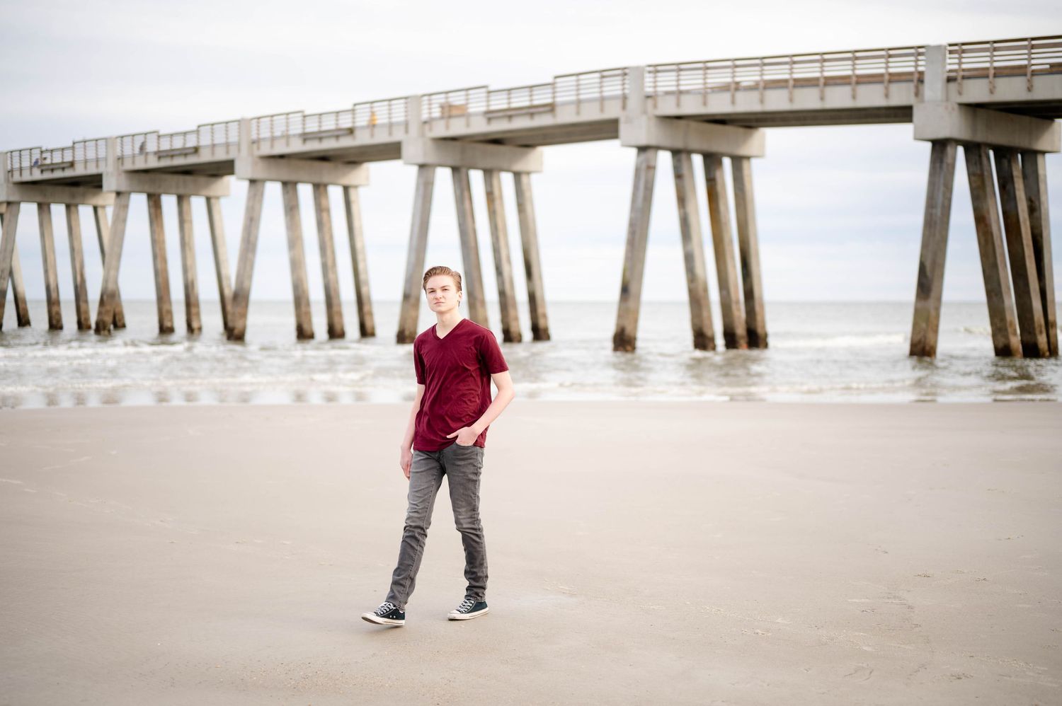 A person in a burgundy sweater walks on an empty beach near a wooden pier on an overcast day.