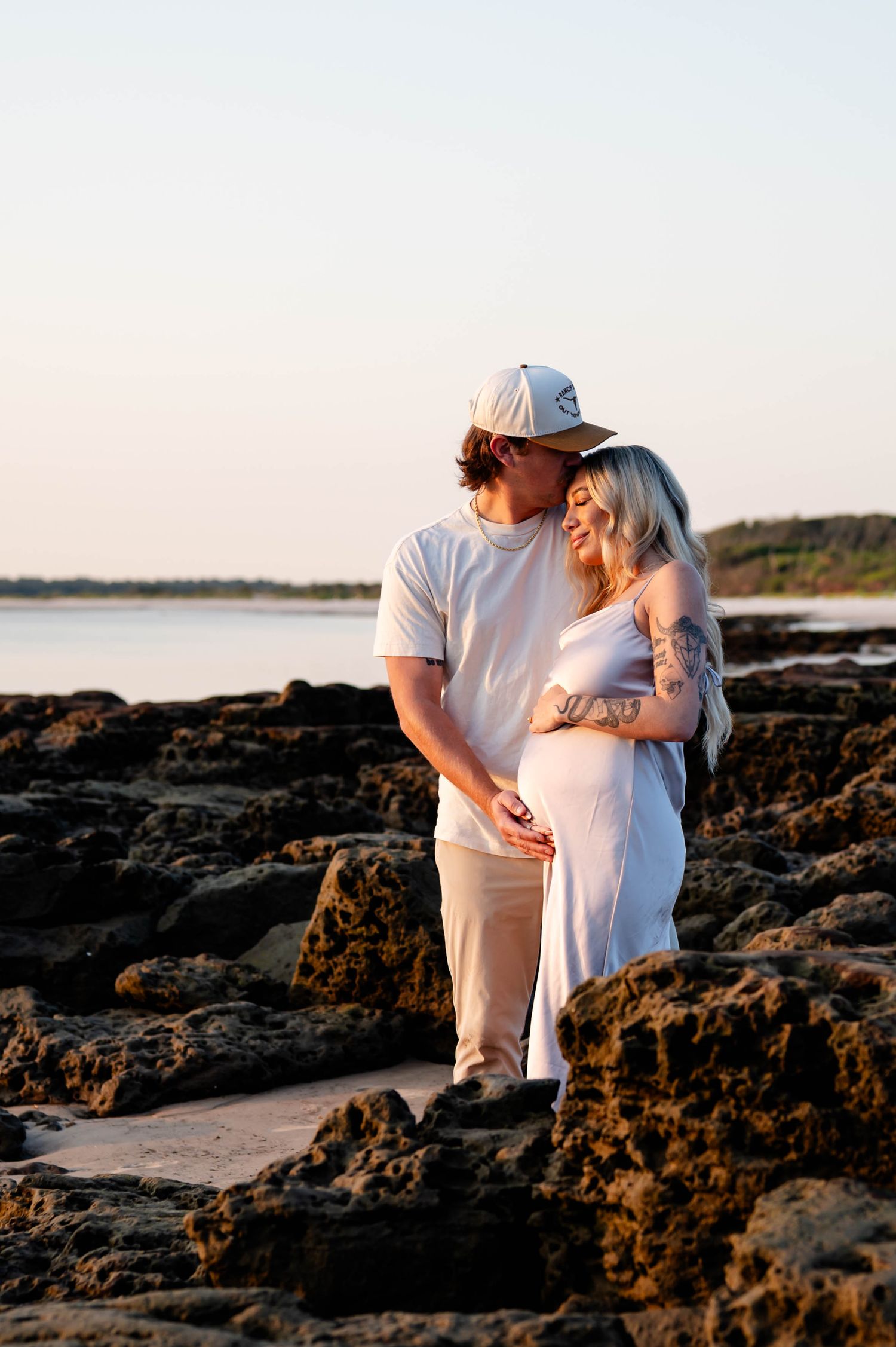 Expectant couple embracing on rocky shore during golden hour.