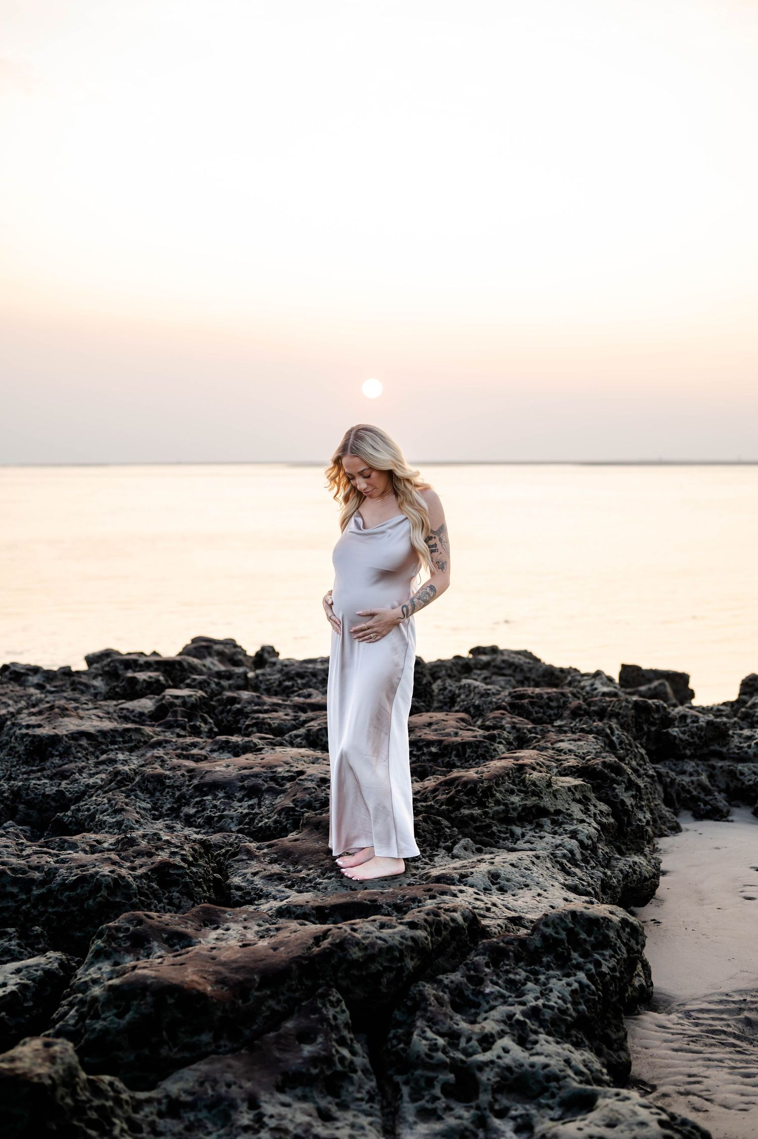 Expectant mother in flowing dress stands on rocky beach at sunrise on Black Rock Beach in Jacksonville - a maternity photoshoot moment.