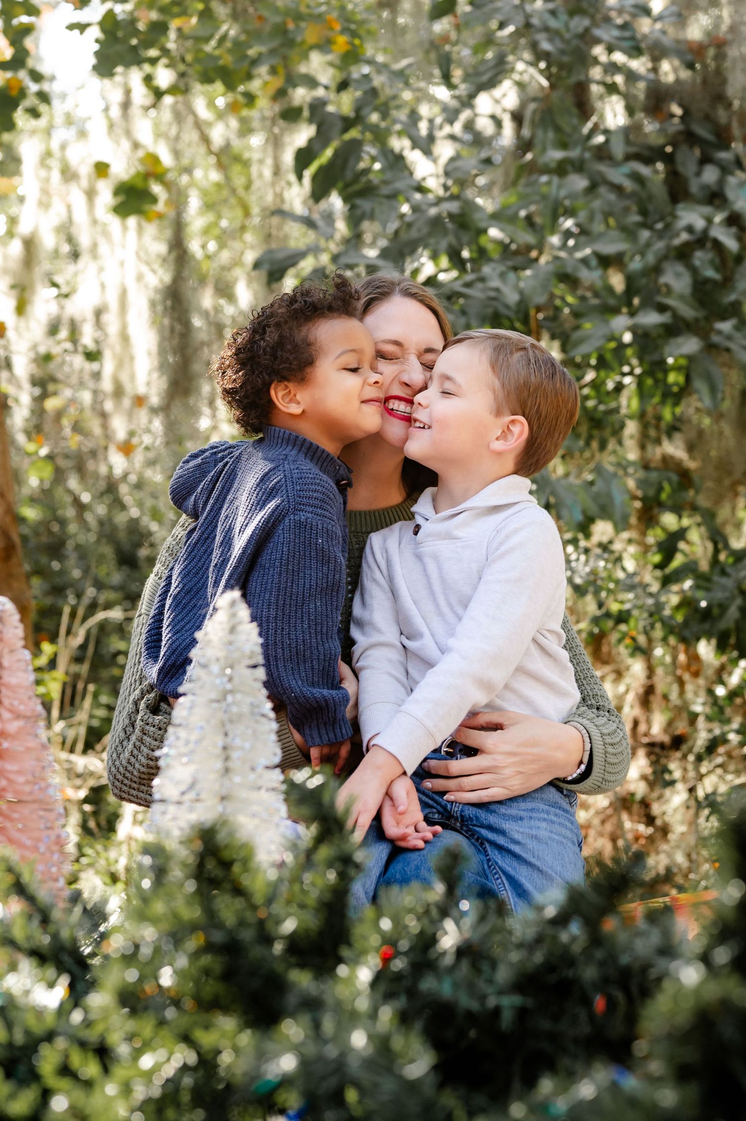Two young children share a sweet moment outdoors in a garden setting with natural sunlight filtering through trees.