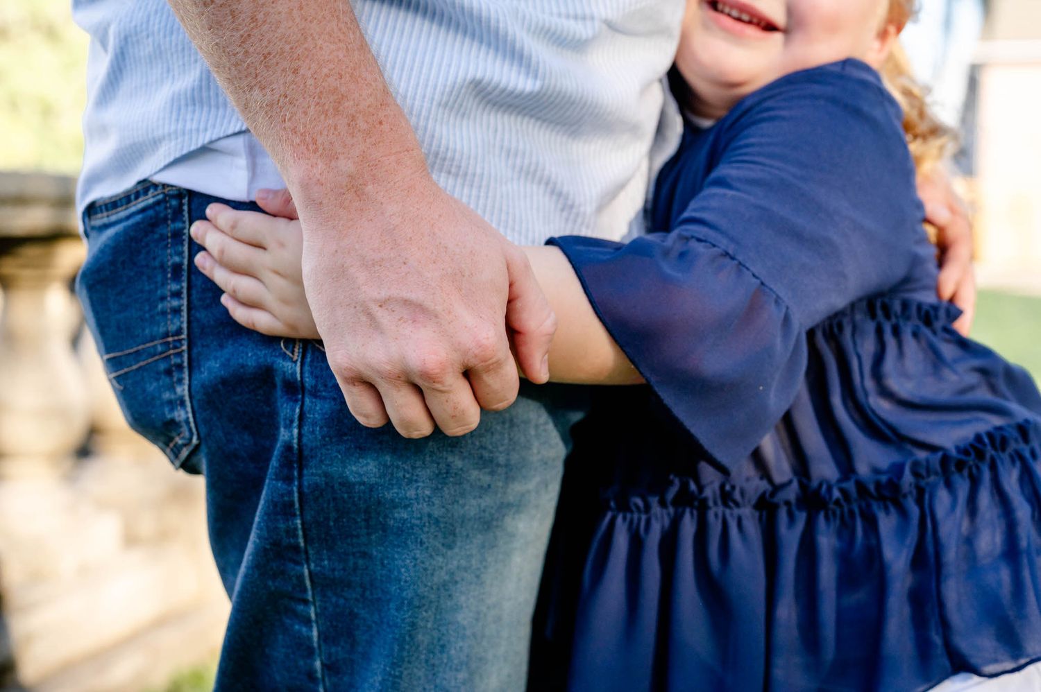 A child in a navy blue dress holds onto an adult's hand and leg while standing outside.