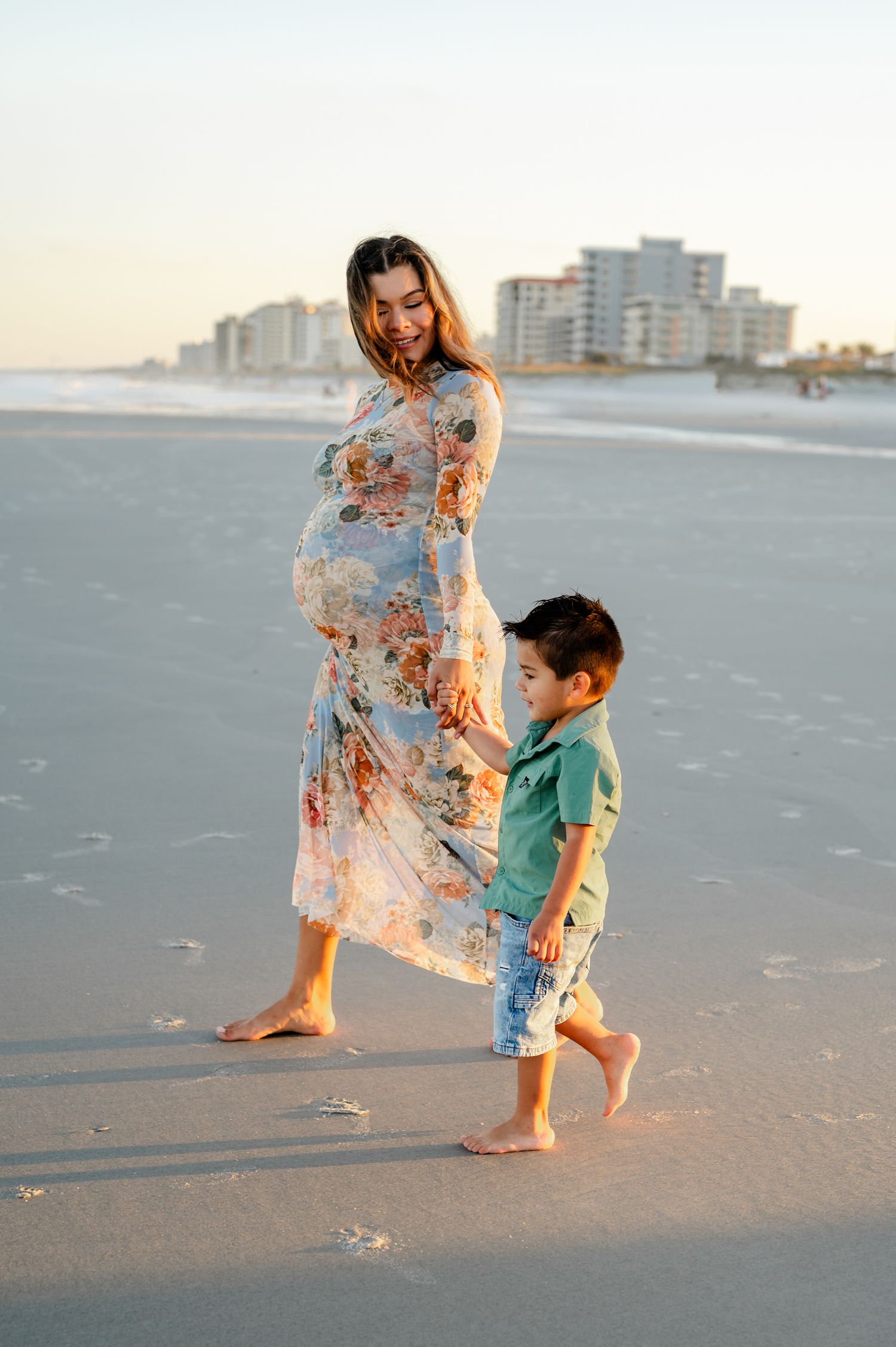 A pregnant woman in a floral dress walks along the beach at sunset with her little boy in a teal shirt during a maternity photoshoot.