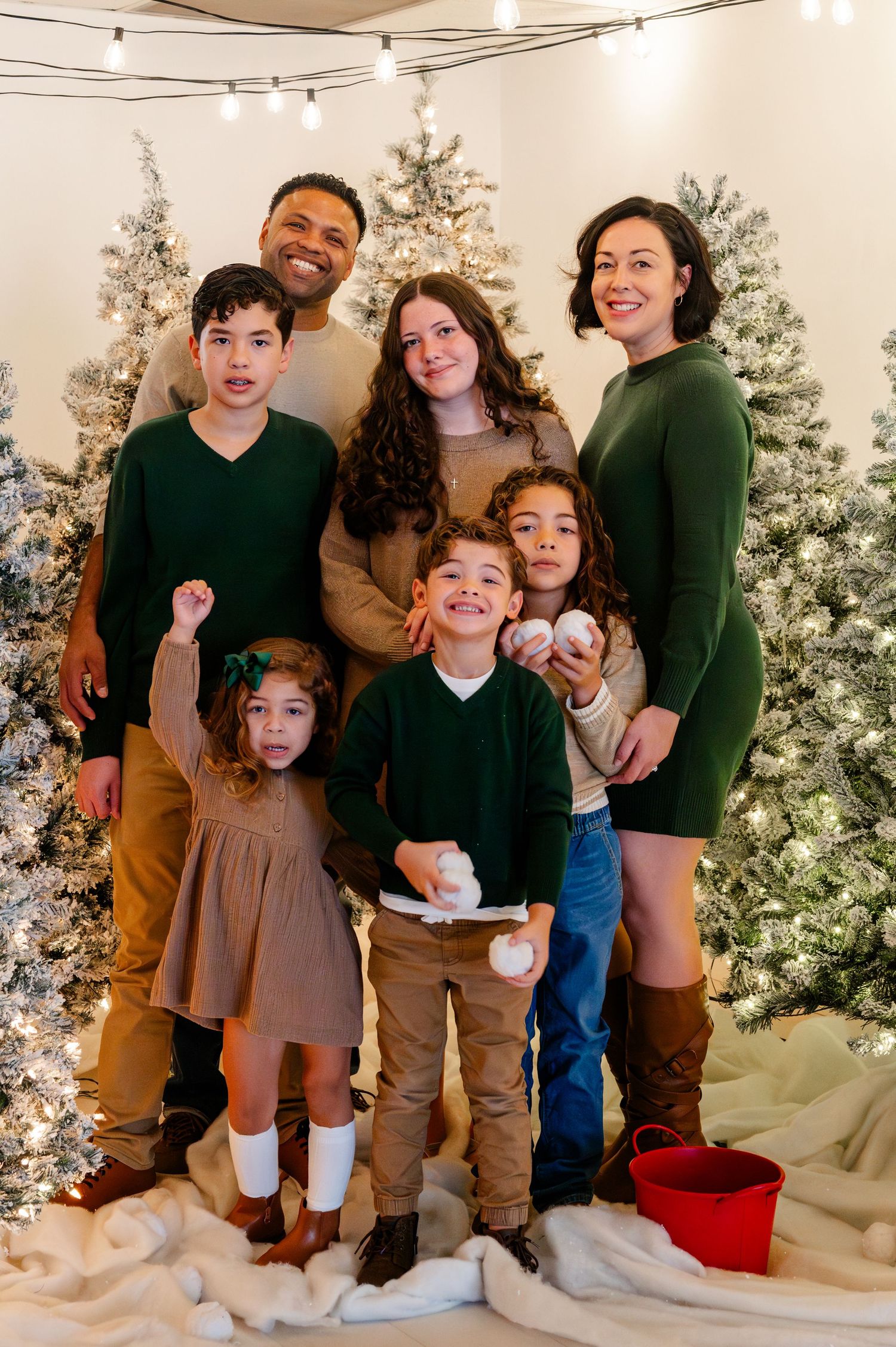Family portrait in coordinated earth-tone outfits posing in front of snow-covered Christmas trees.
