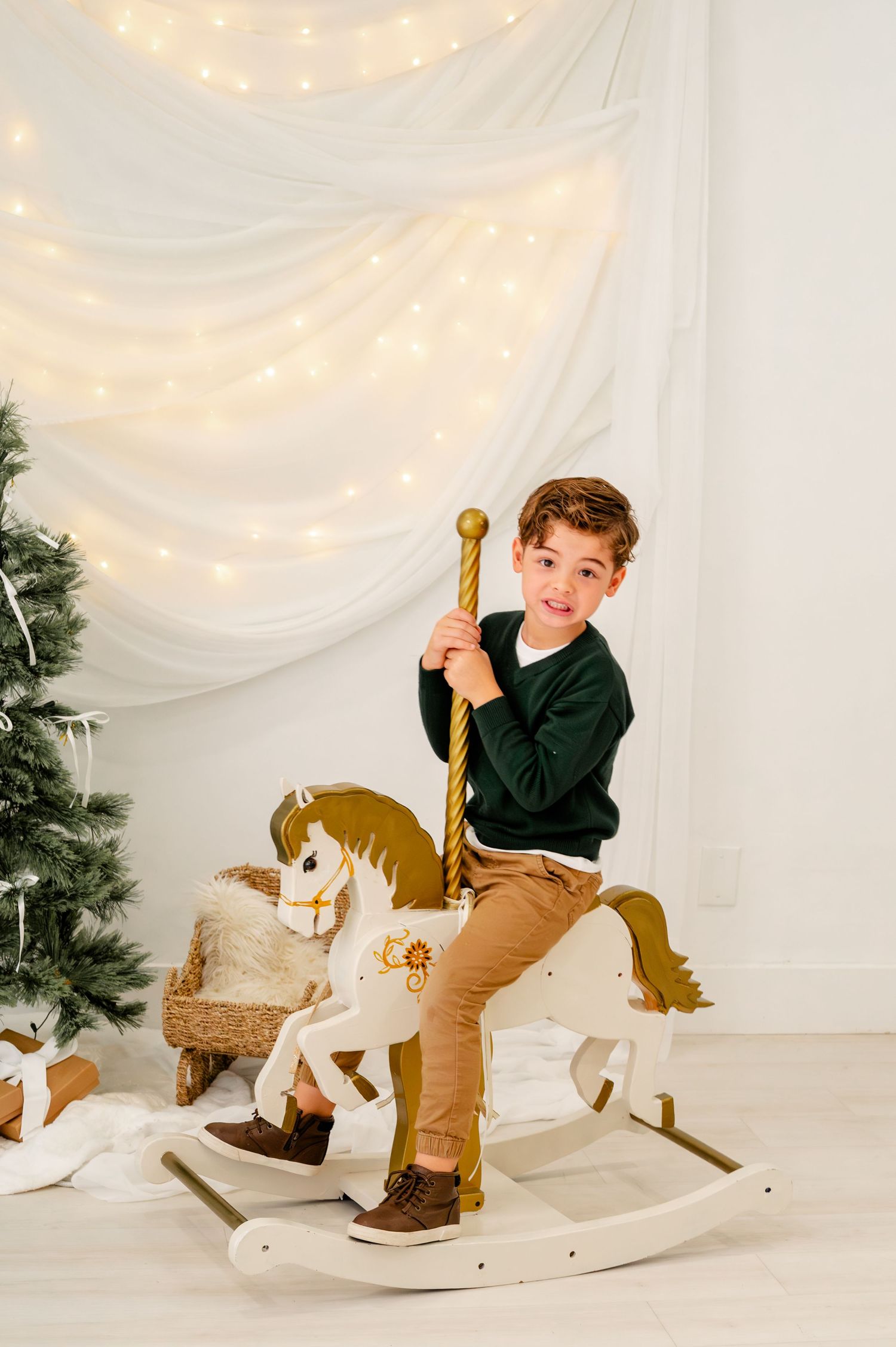 A child in a black sweater and khaki pants plays on a white rocking horse against a white backdrop with holiday decor.