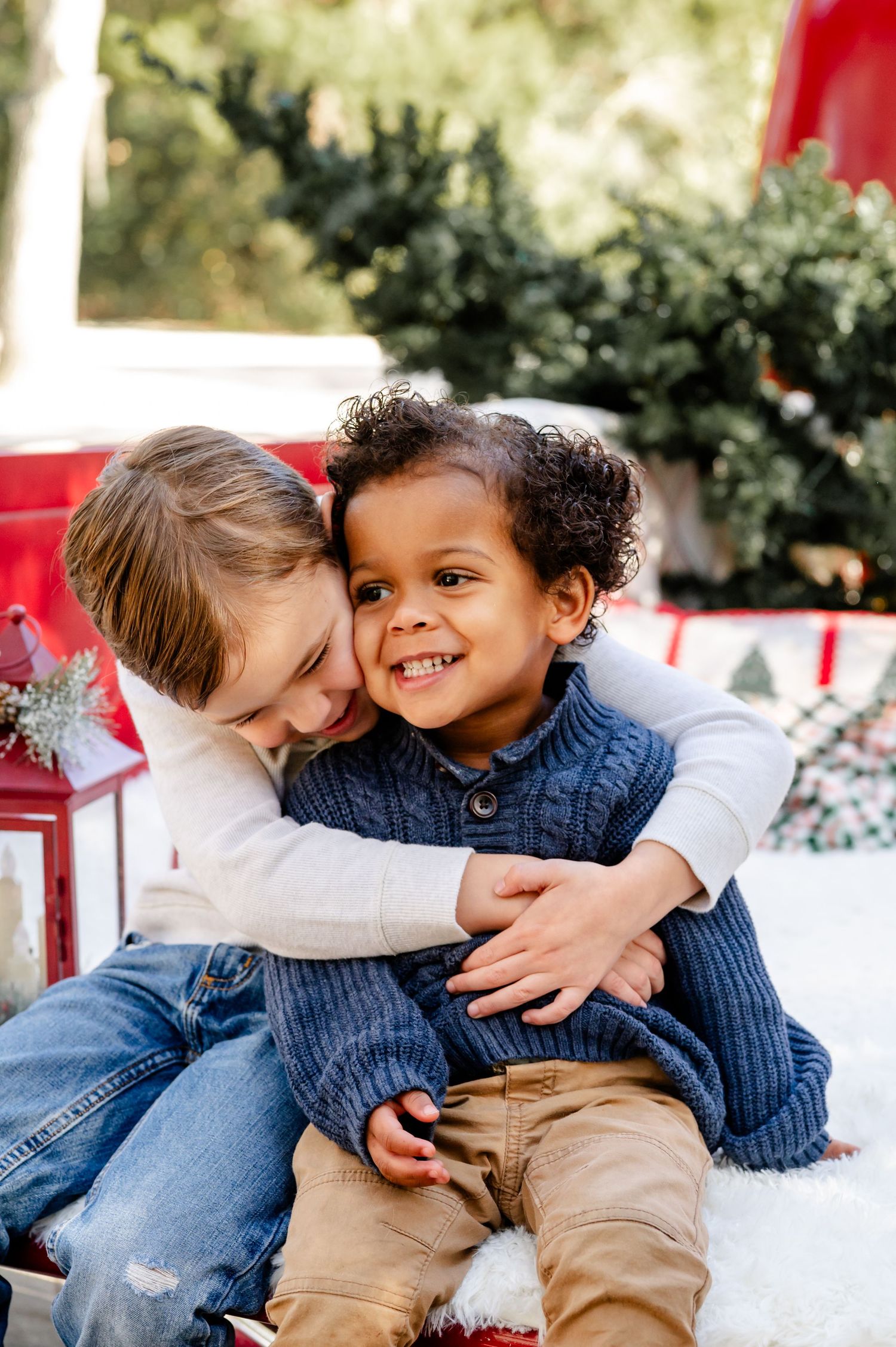 Two children share a sweet embrace while sitting together in a winter holiday setting with red and white decor.