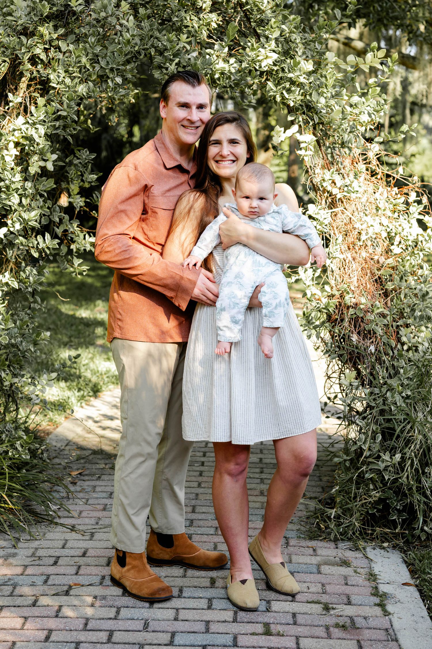 A young family poses together on a brick pathway surrounded by lush greenery on a sunny day.