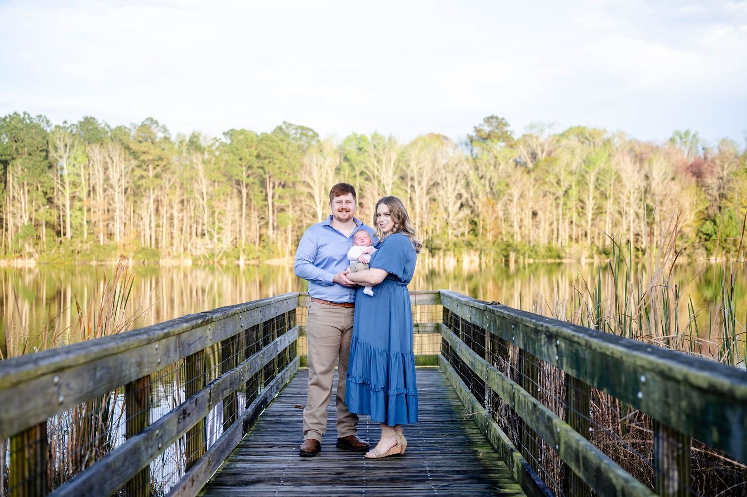 Couple standing together holding baby on wooden boardwalk over lake with cypress trees.