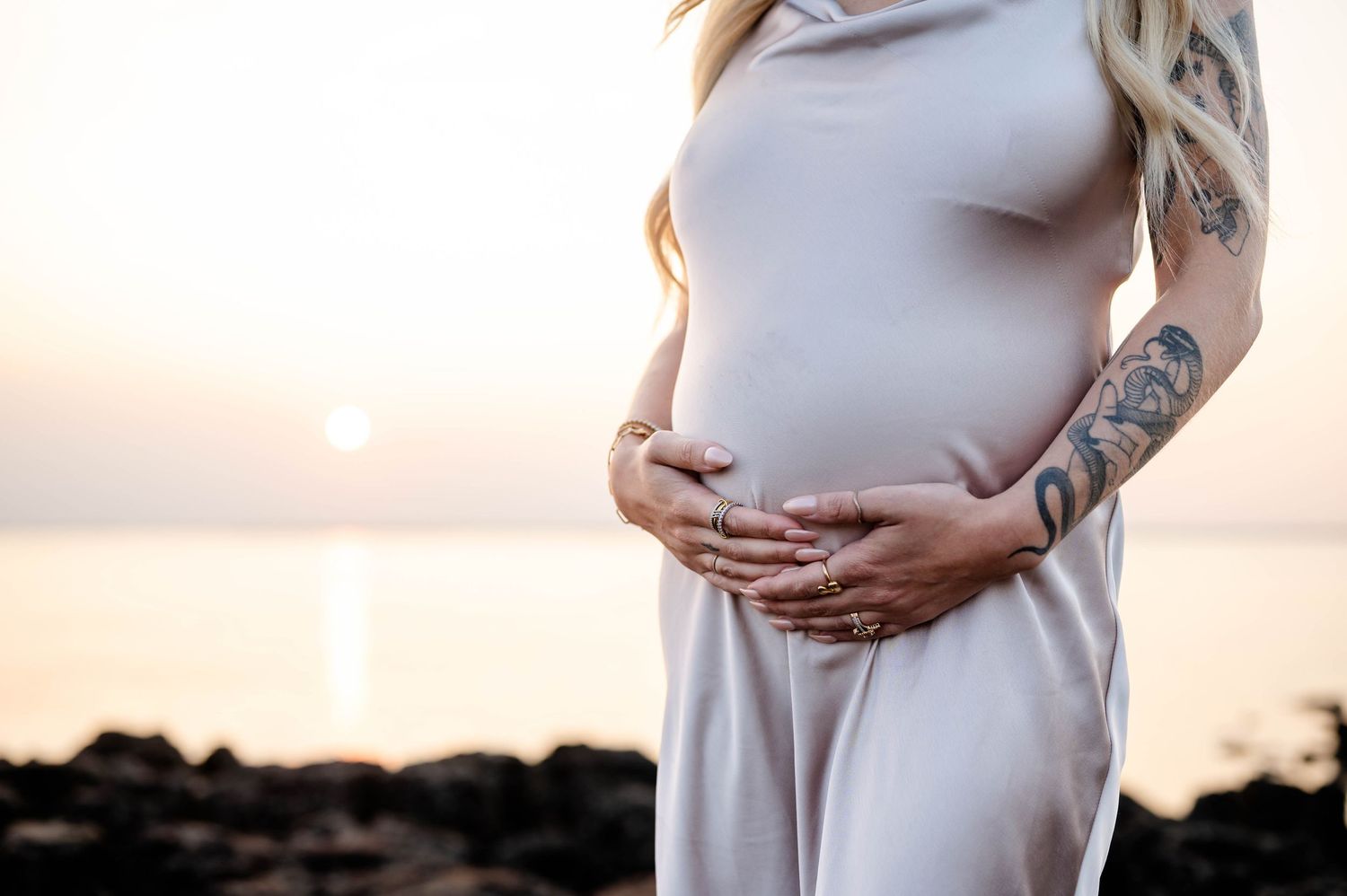 Side profile of pregnant woman in flowing white dress against sunset beach backdrop.