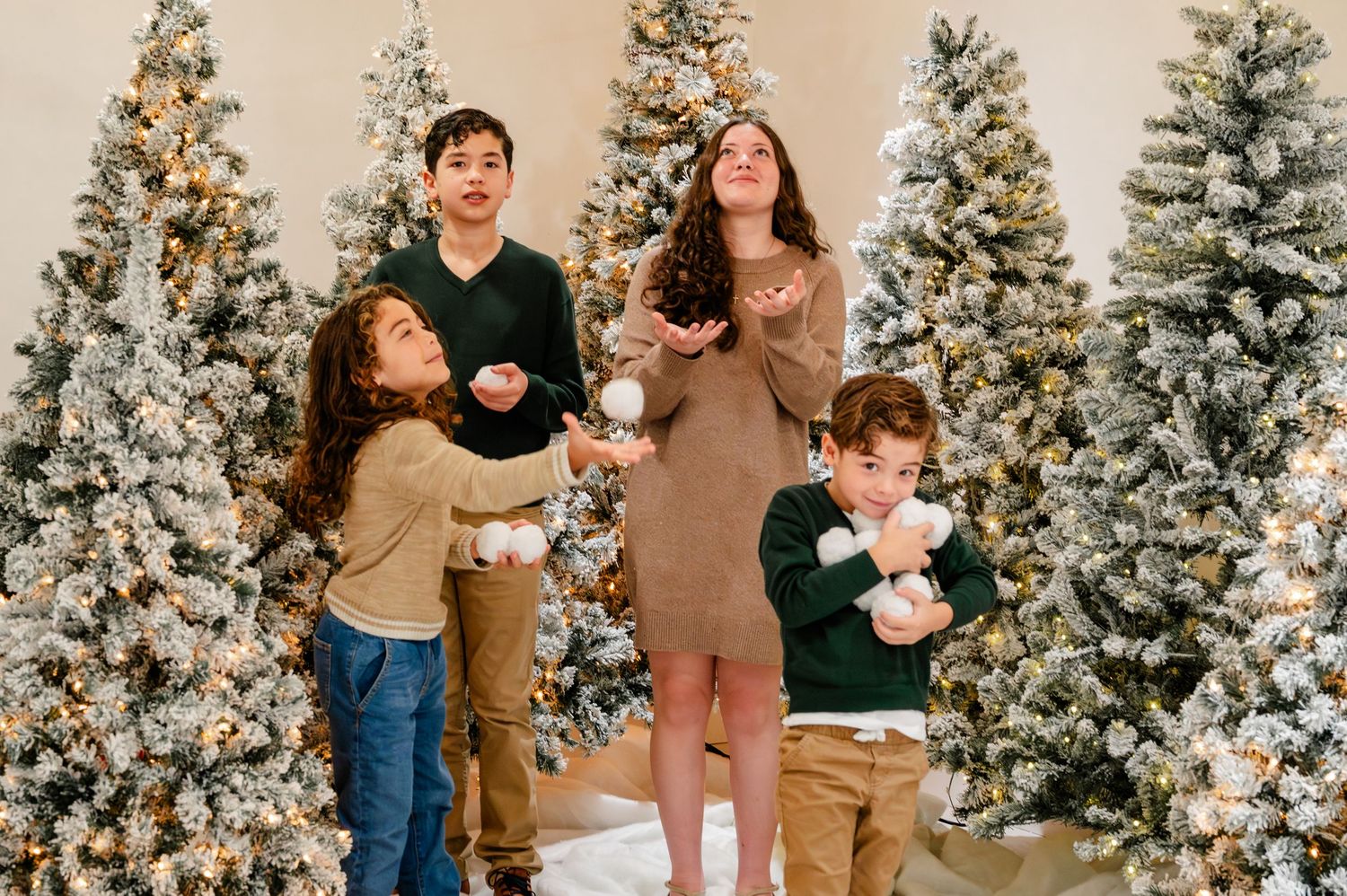Family wearing coordinated brown and black outfits poses in front of snow-covered Christmas trees for holiday photos.
