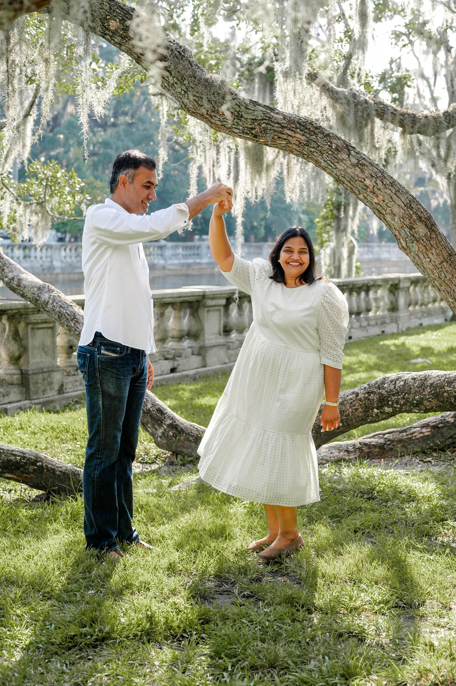 Couple in white and denim dance playfully under moss-draped trees in a romantic outdoor setting.