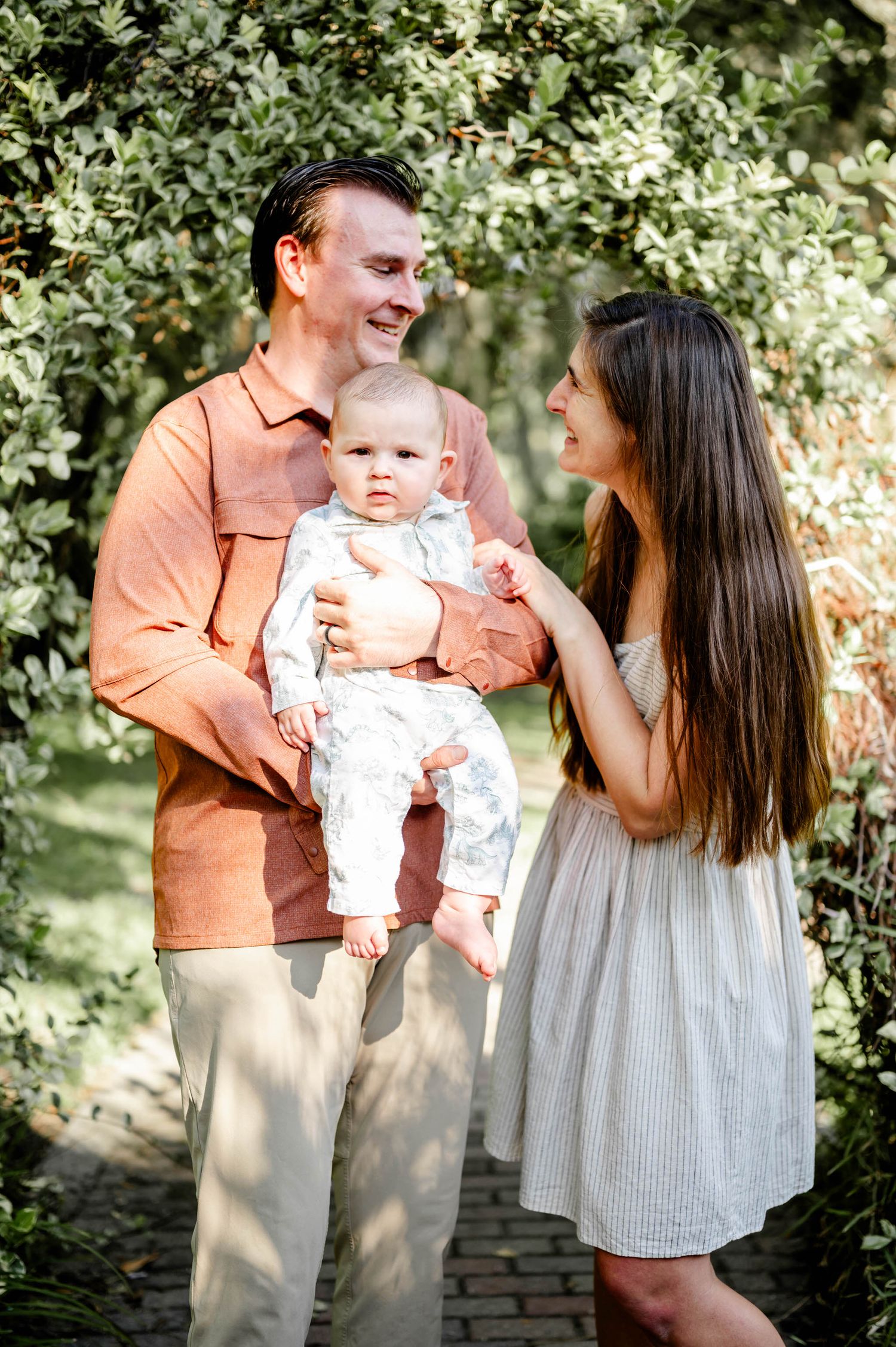 A family shares tender moments together in a garden setting with dappled sunlight filtering through the trees.