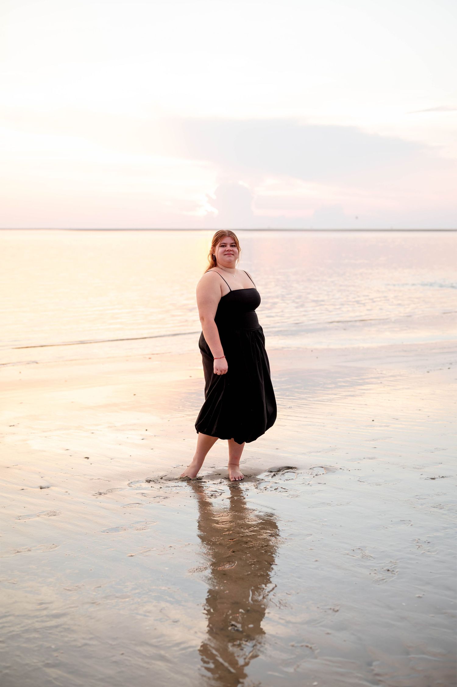 A person wearing a black dress stands in shallow ocean water during sunset at the beach.