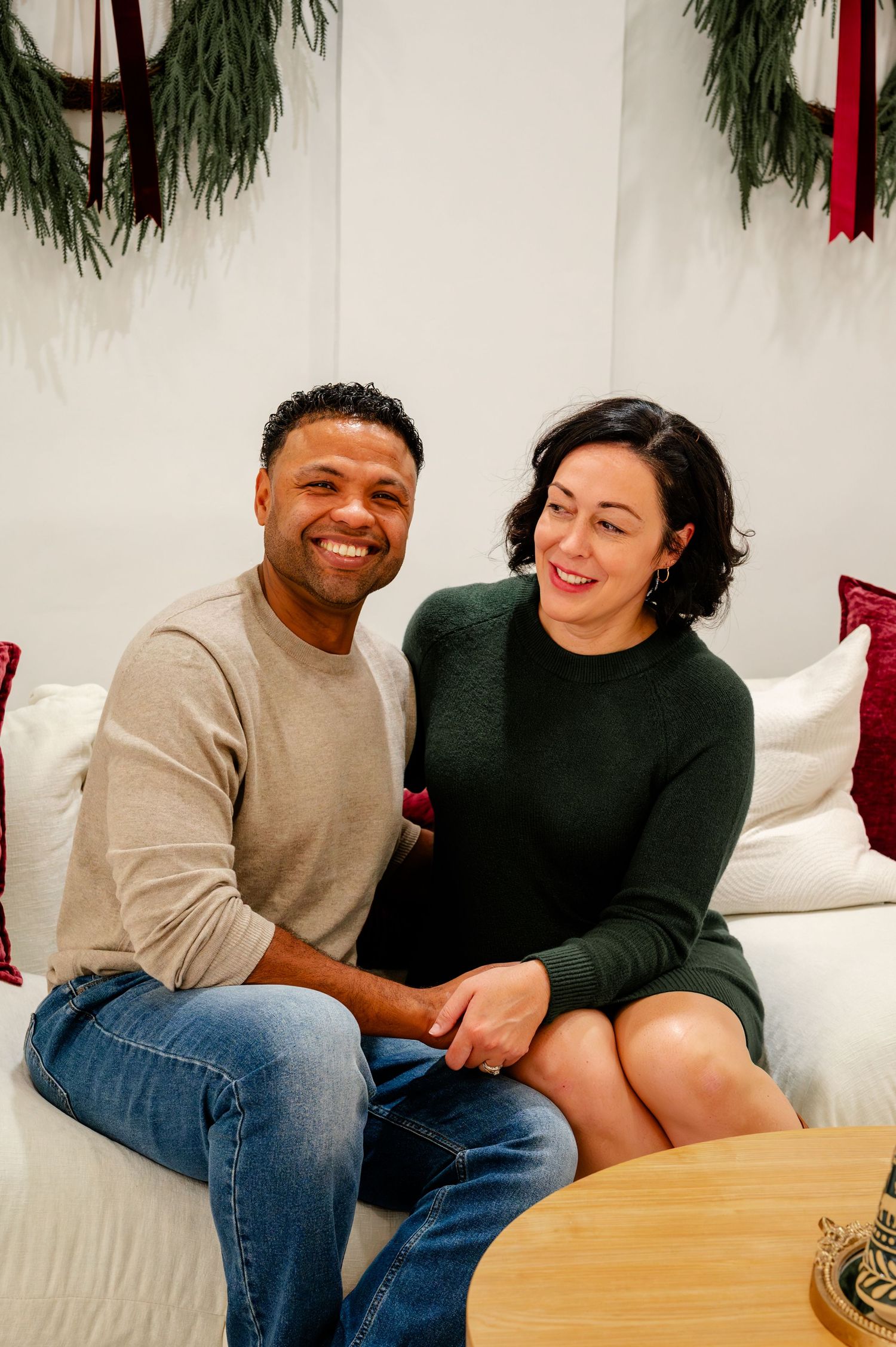 A couple sits together on a white couch, sharing laughter and intimate moments in a festive holiday setting.