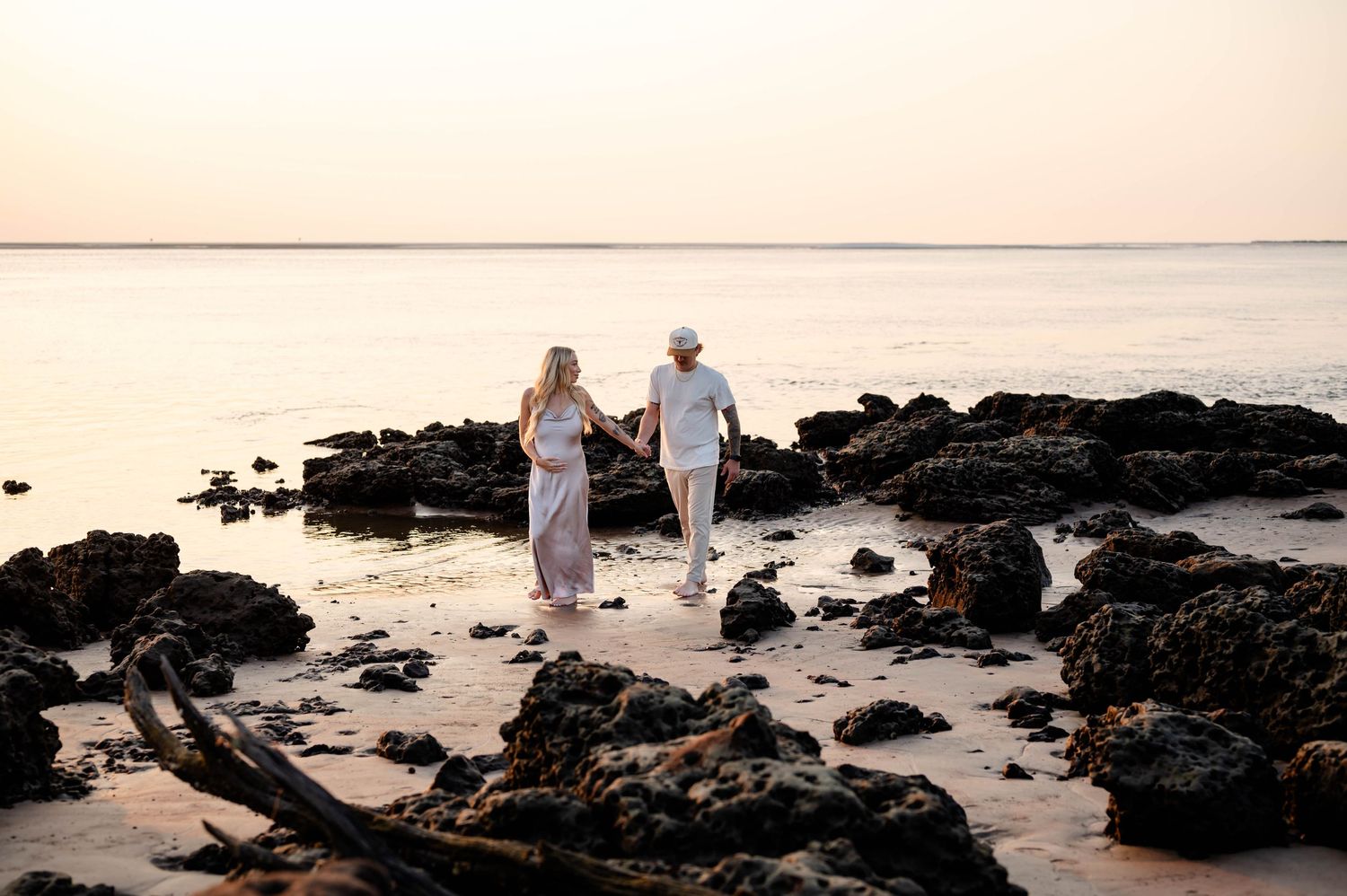 Couple walking along rocky beach shoreline at sunset wearing light colored clothing.