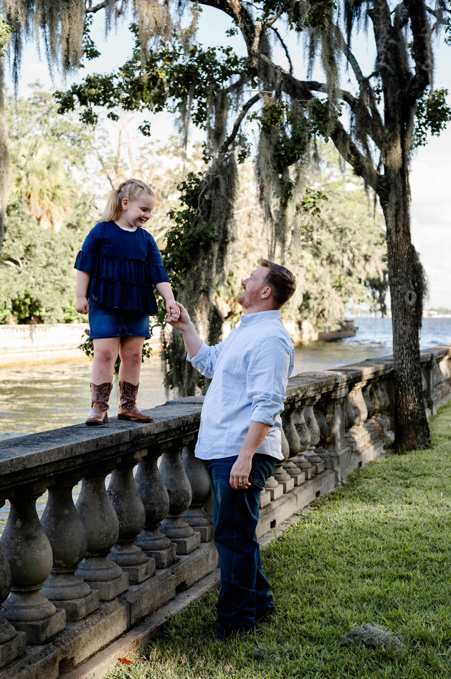 Two people interact playfully on a stone balustrade in a scenic garden setting with Spanish moss hanging from trees.