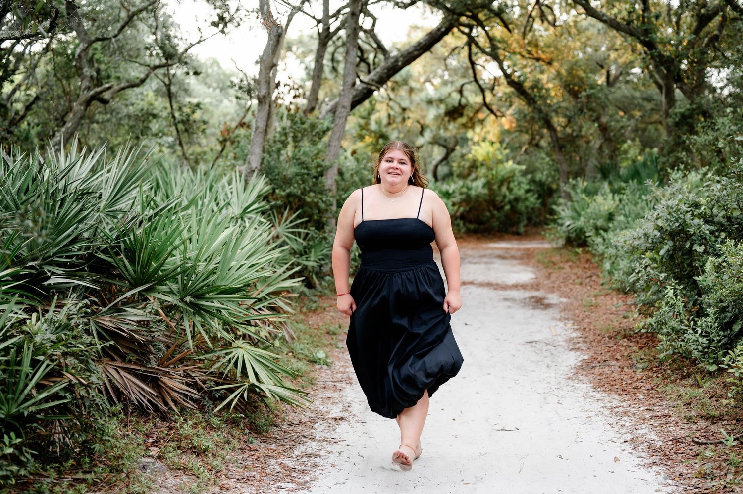 A person in formal attire walks along a scenic wooded path among palm trees and greenery.