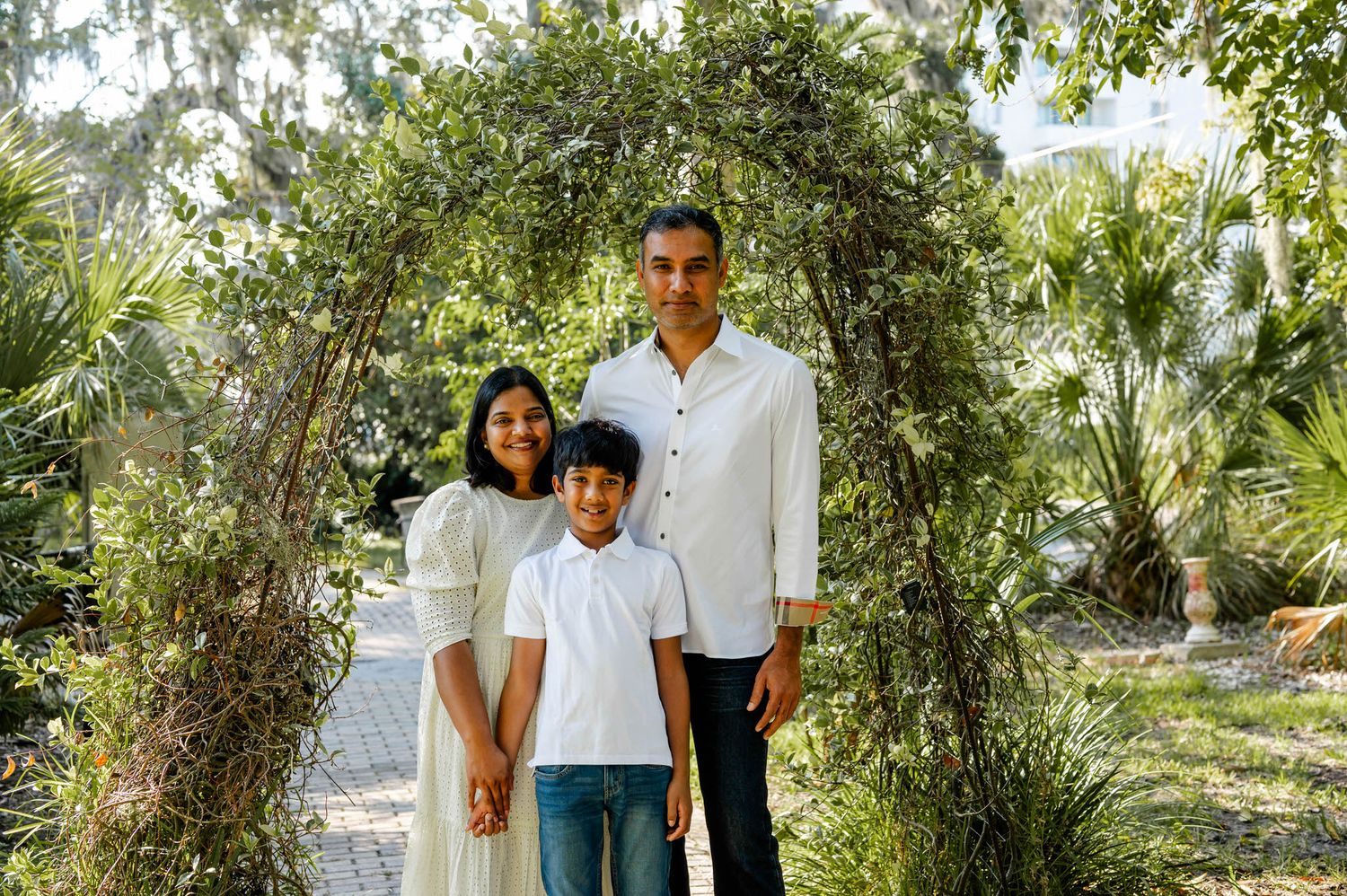A couple stands together in a lush outdoor garden setting with green foliage and trees in the background.