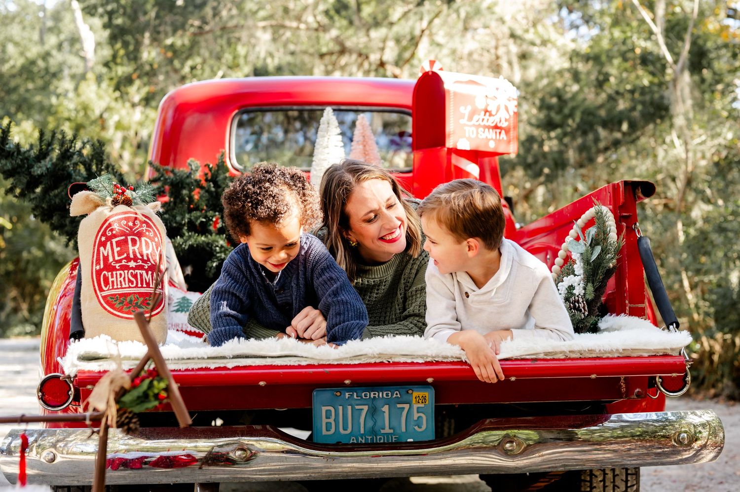 Three people laughing together while sitting in the bed of a vintage red pickup truck on a sunny day.