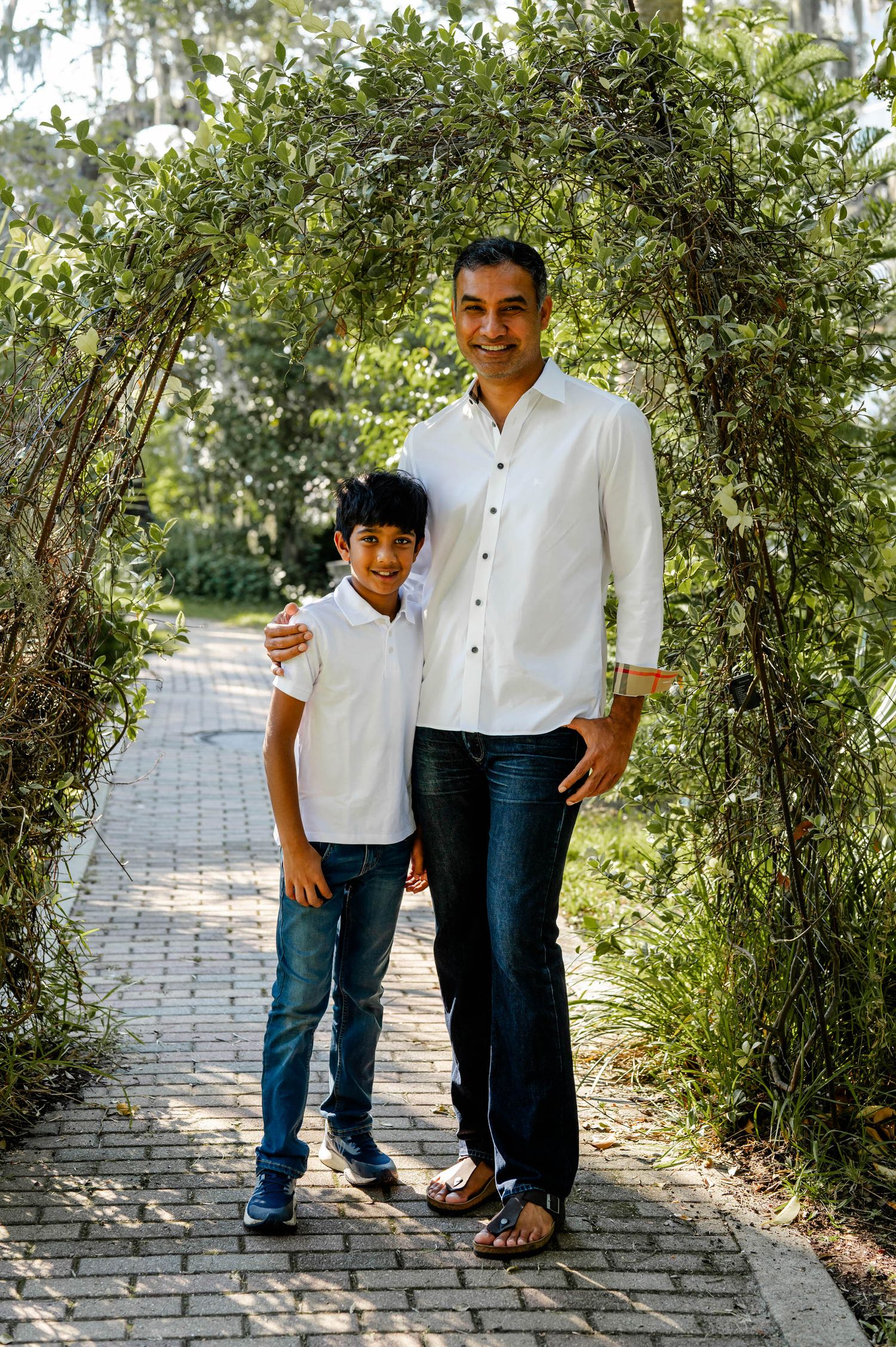 A parent and child wearing matching white button-down shirts and jeans stand together under a garden archway.