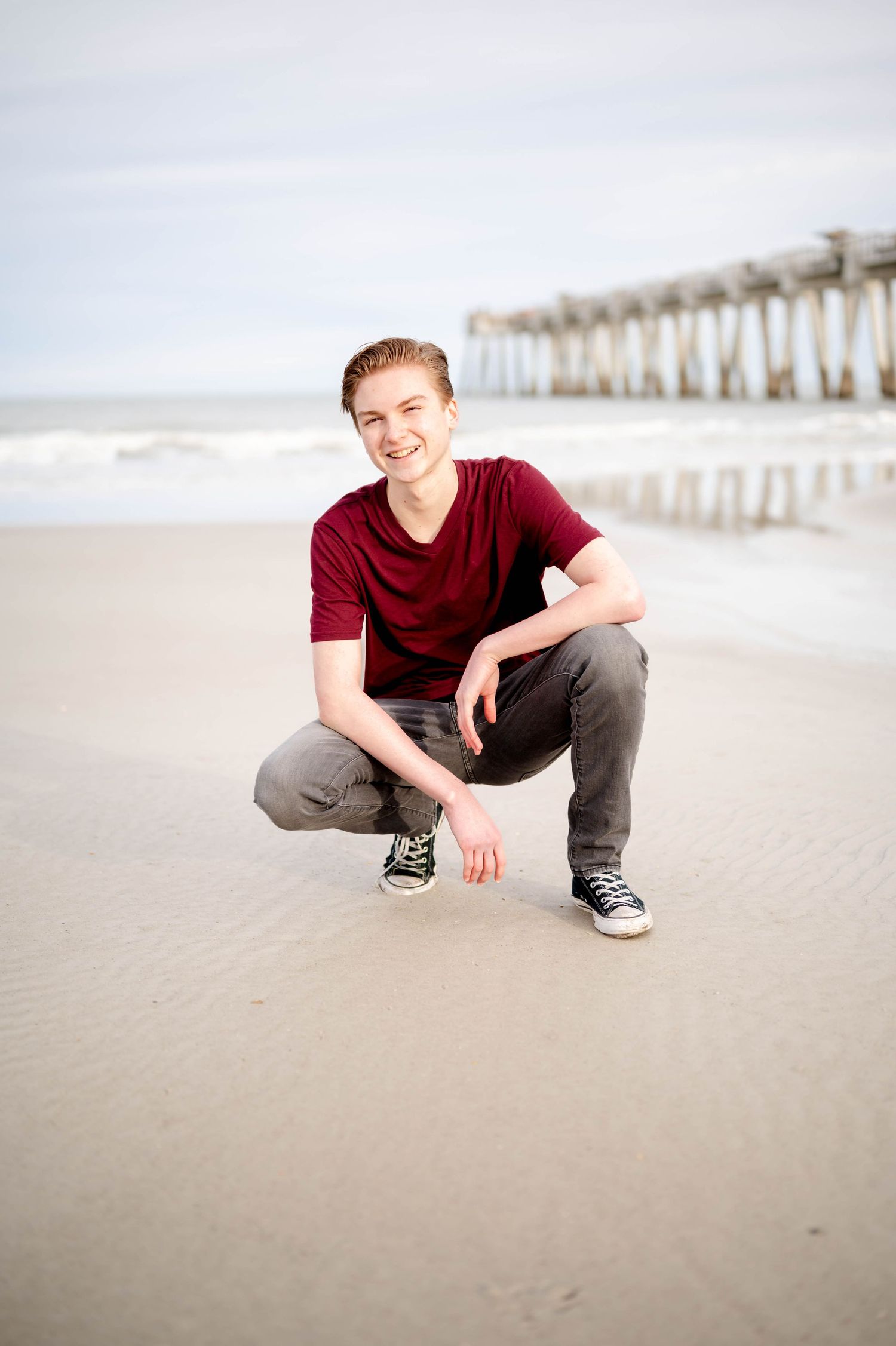 Person in red t-shirt and gray pants poses on sandy beach with pier visible in background.