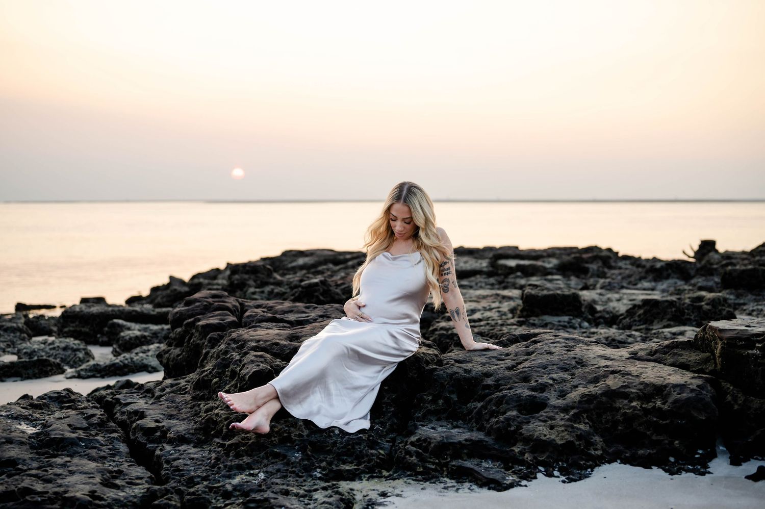 A figure in a flowing white dress sits on dark coastal rocks at sunset as ocean waves meet the shore in the background.