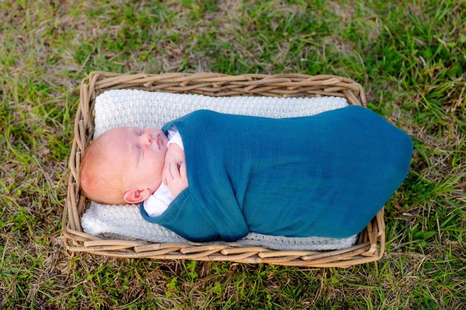 Sleeping newborn in Jacksonville FL photographed while wrapped in blue swaddle blanket resting in wicker basket outdoors.