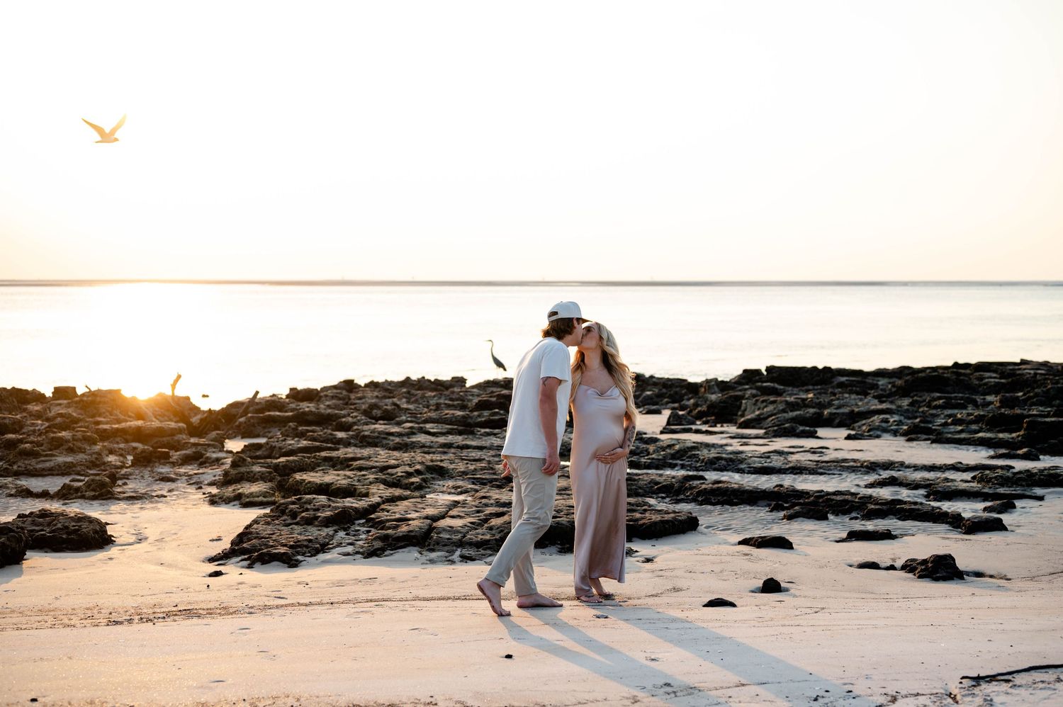 Wide landscape shot of a beach at sunset with dark volcanic rocks and two distant figures.
