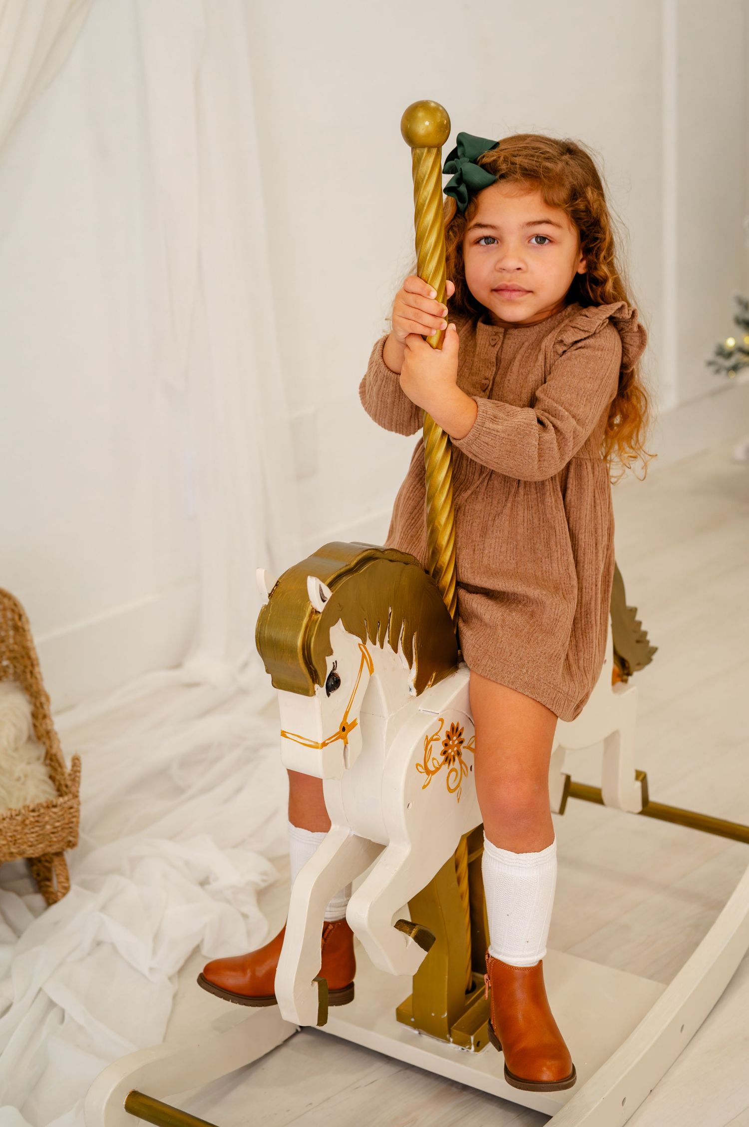 A young child in a brown sweater and white boots poses playfully with a gold Christmas ornament.